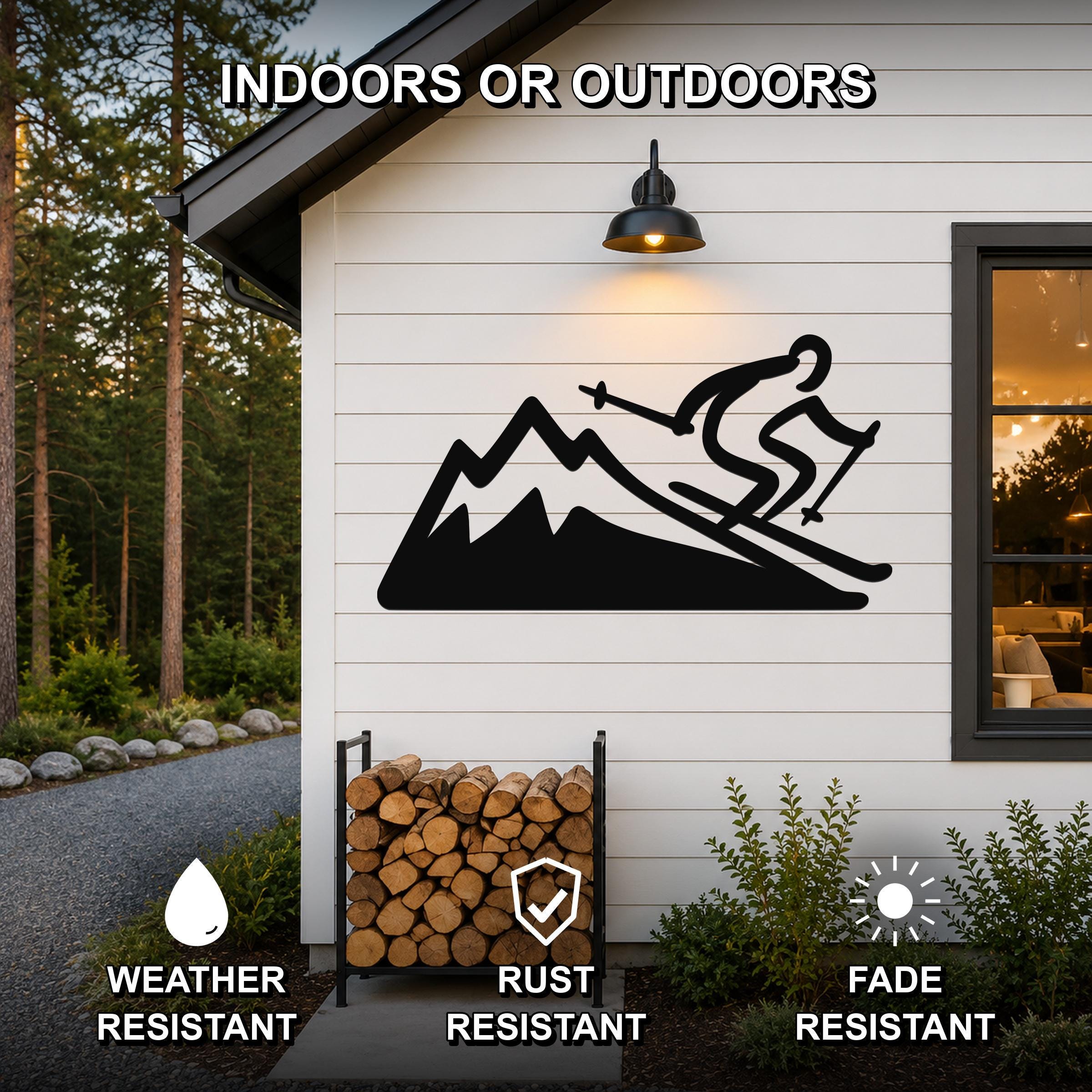 Black metal wall sculpture of a skier on a mountain, mounted on a white siding wall outside a home, with a firewood rack below and a light fixture above.