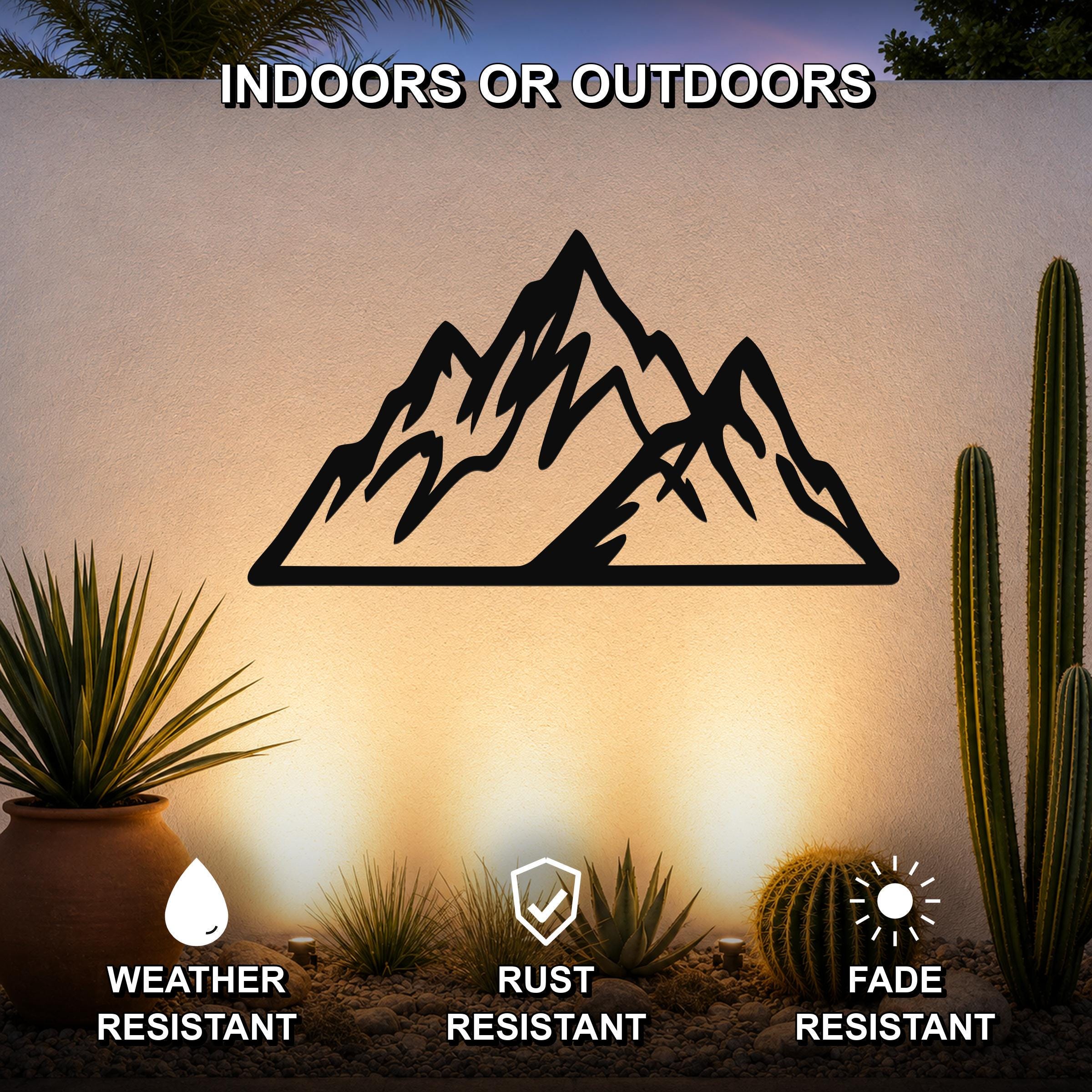 Black metal mountain wall art with layered peak cutouts, mounted on an outdoor wall above a desert garden with cacti and pebbles.