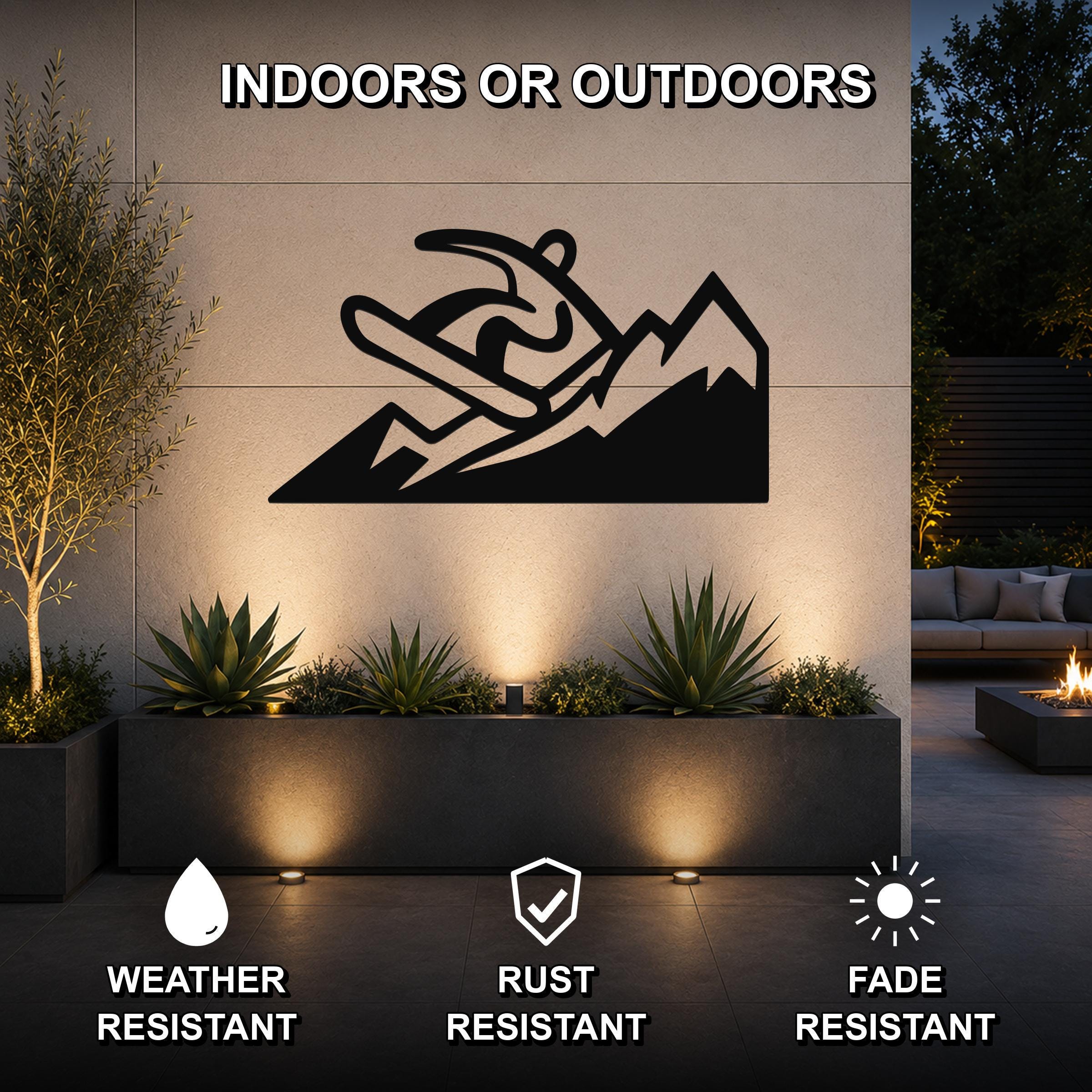 Black metal wall art featuring a snowboarder silhouette against mountain peaks, mounted on an outdoor wall above a planter in a modern patio setting.