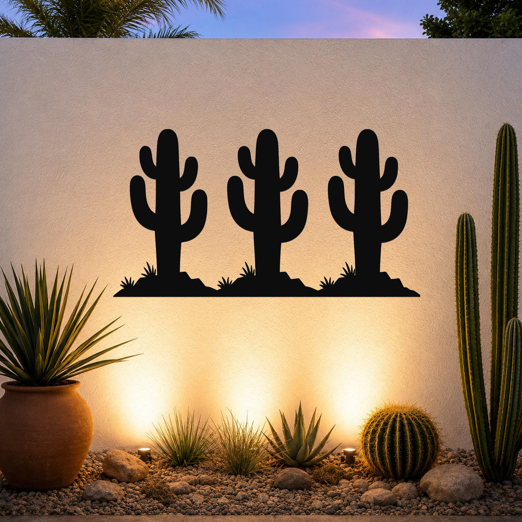 Black metal wall art featuring a trio of saguaro cactus silhouettes with cutout arms and bases, mounted on an outdoor wall above a desert garden with potted plants and ground lights.