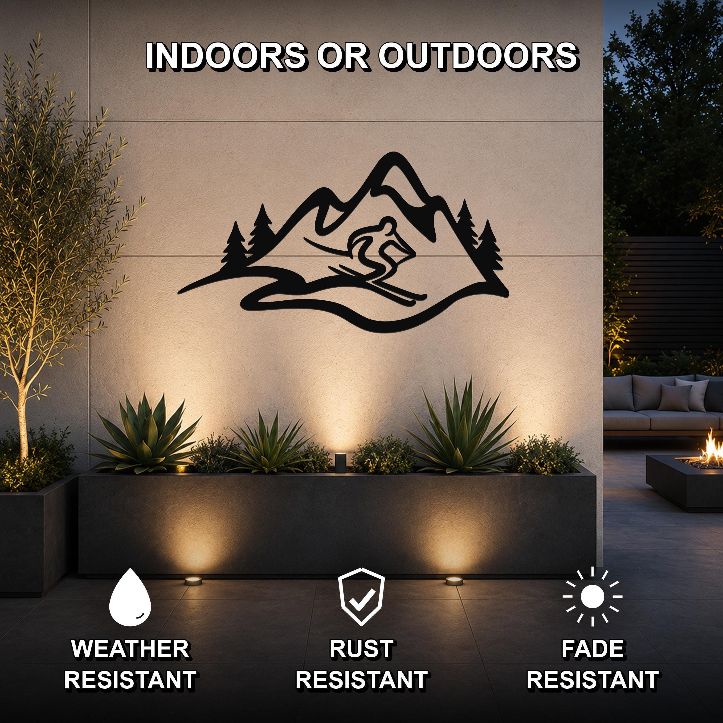 Black metal wall art featuring a skier silhouette within a mountain range and pine trees, mounted on an outdoor wall above a planter with uplighting.