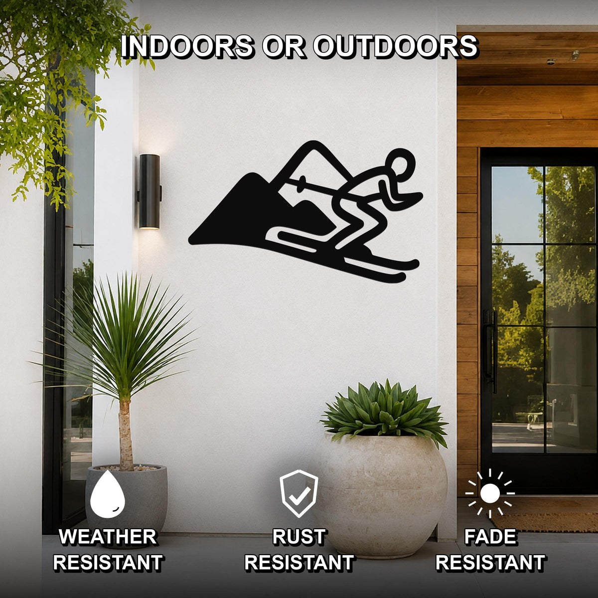 Black metal skier and mountain wall art silhouette mounted on a white exterior wall beside a glass door and potted plants, with text indicating it is weather, rust, and fade resistant.