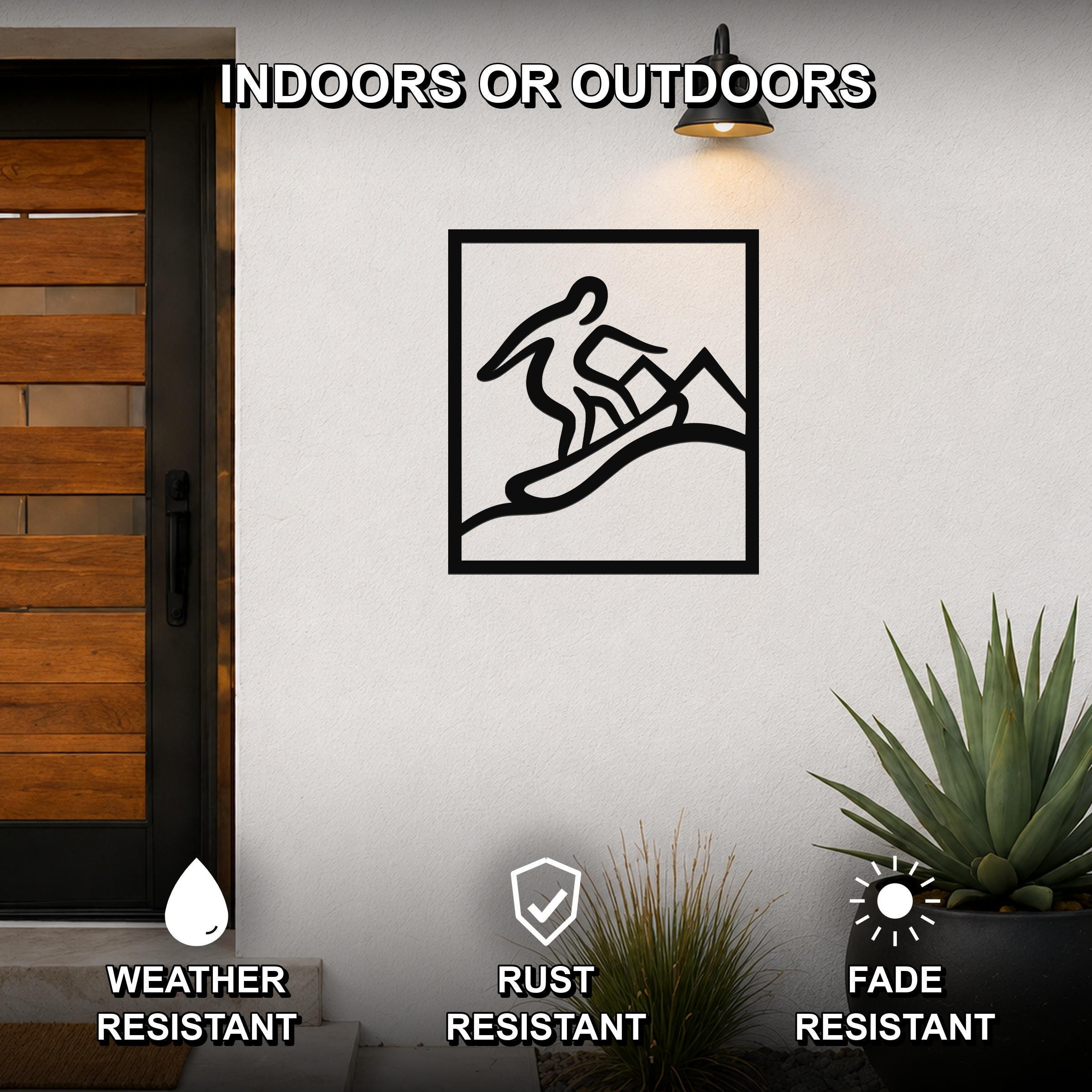Black metal wall art featuring a minimalist silhouette of a snowboarder in motion, mounted on an outdoor wall beside a wooden door and potted plants.