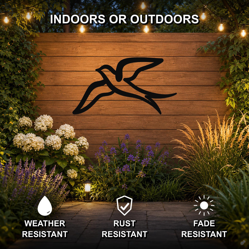 Black metal swallow bird wall art silhouette mounted on a wooden outdoor wall, displayed above a garden bed with flowers and string lights, weather resistant, rust resistant, and fade resistant.