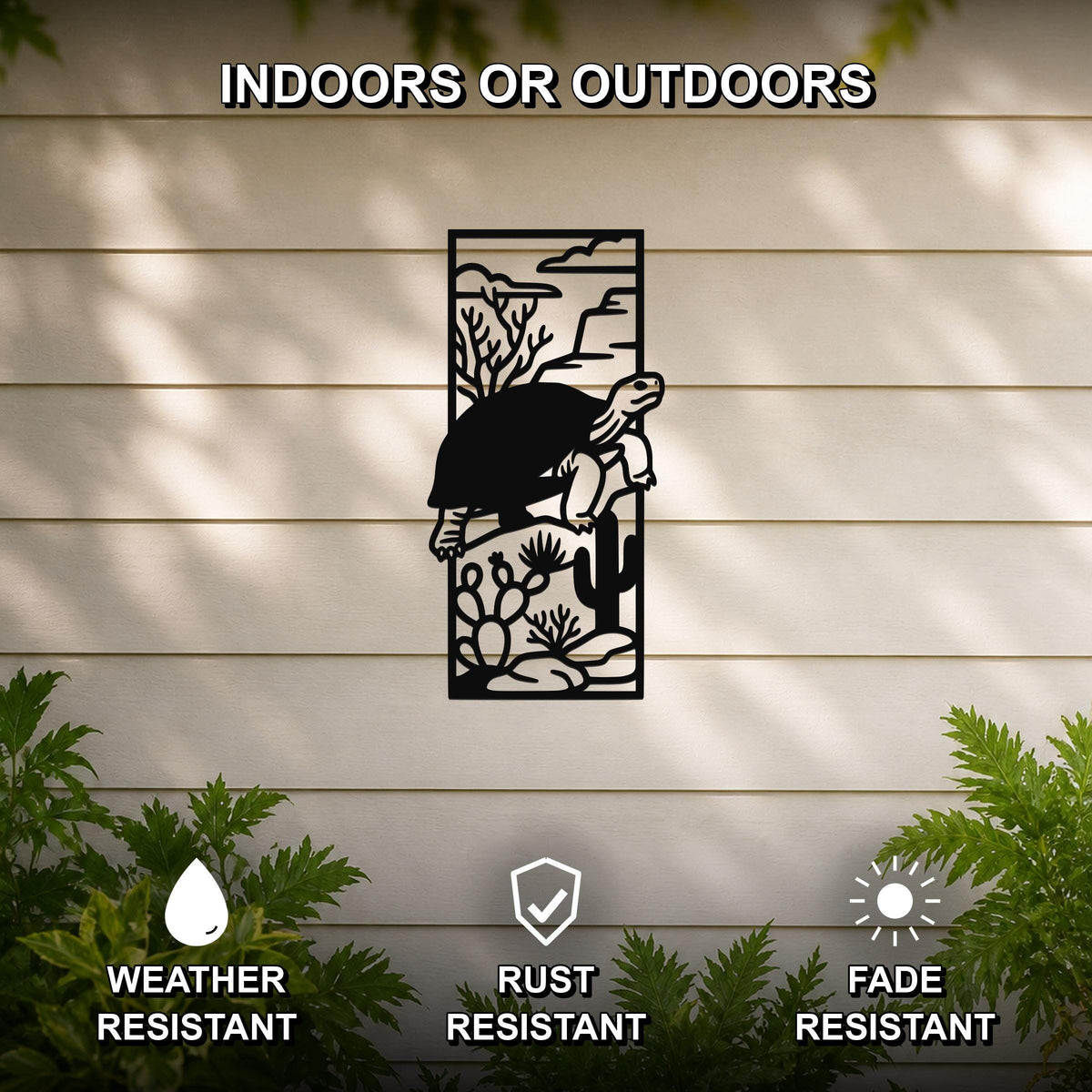 Black metal wall art featuring a tortoise silhouette climbing over cacti and desert plants, mounted on a light-colored outdoor wall with greenery below.