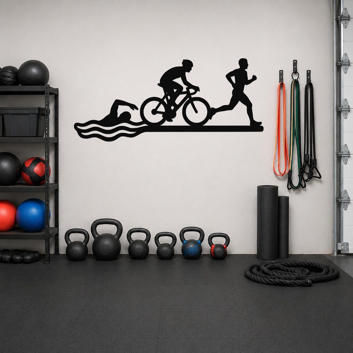 Black metal wall art cutout of a triathlon scene with swim, bike, and run silhouettes, mounted on a gym wall above a row of kettlebells.
