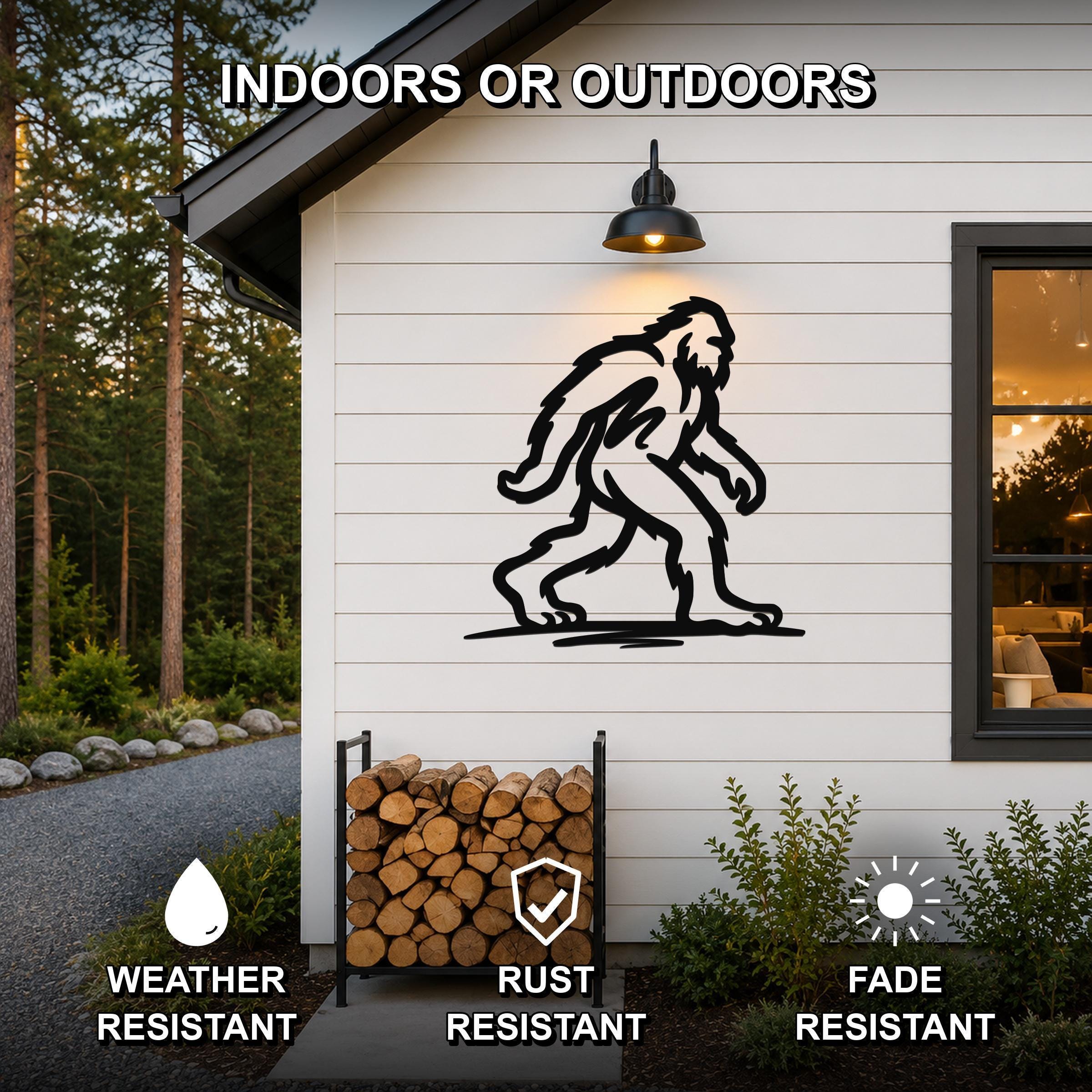Black metal Bigfoot silhouette wall art mounted on a white siding exterior wall above a firewood rack, with text indicating it is weather, rust, and fade resistant for indoor or outdoor use.
