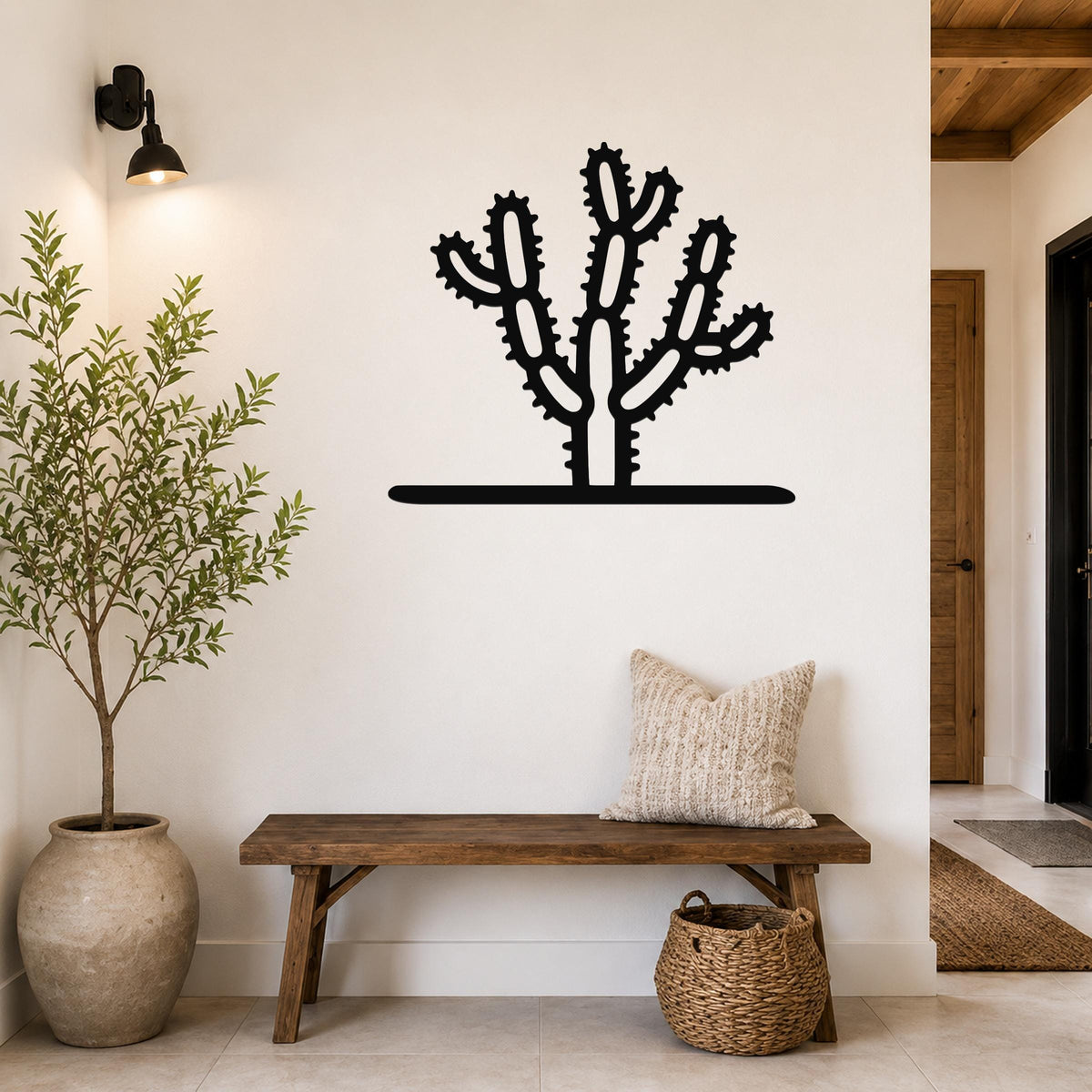 Black metal cholla cactus wall art with spiny branch cutouts, mounted on a white wall above a wooden bench in a entryway.