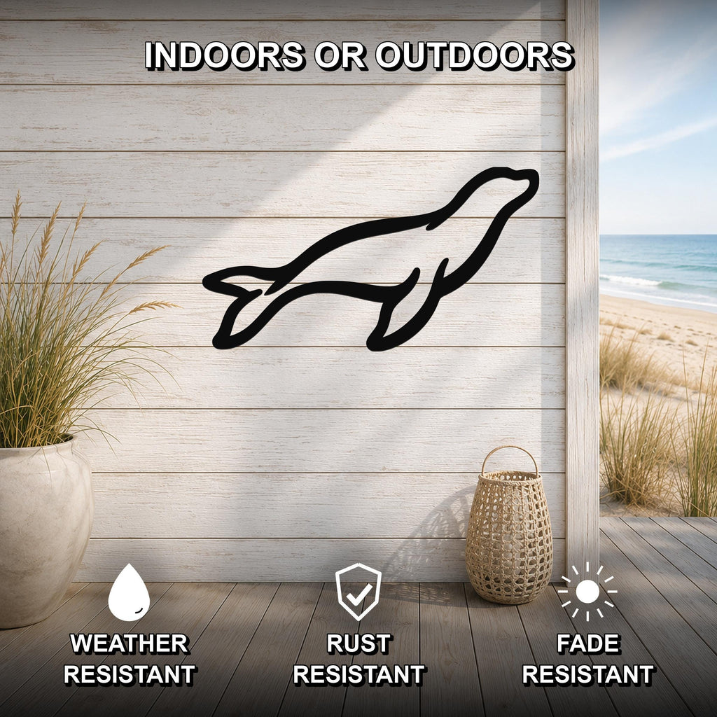 Black metal sea lion silhouette wall art mounted on a light wood plank wall, shown in an indoor-outdoor setting with a beach view, weather resistant, rust resistant, and fade resistant.