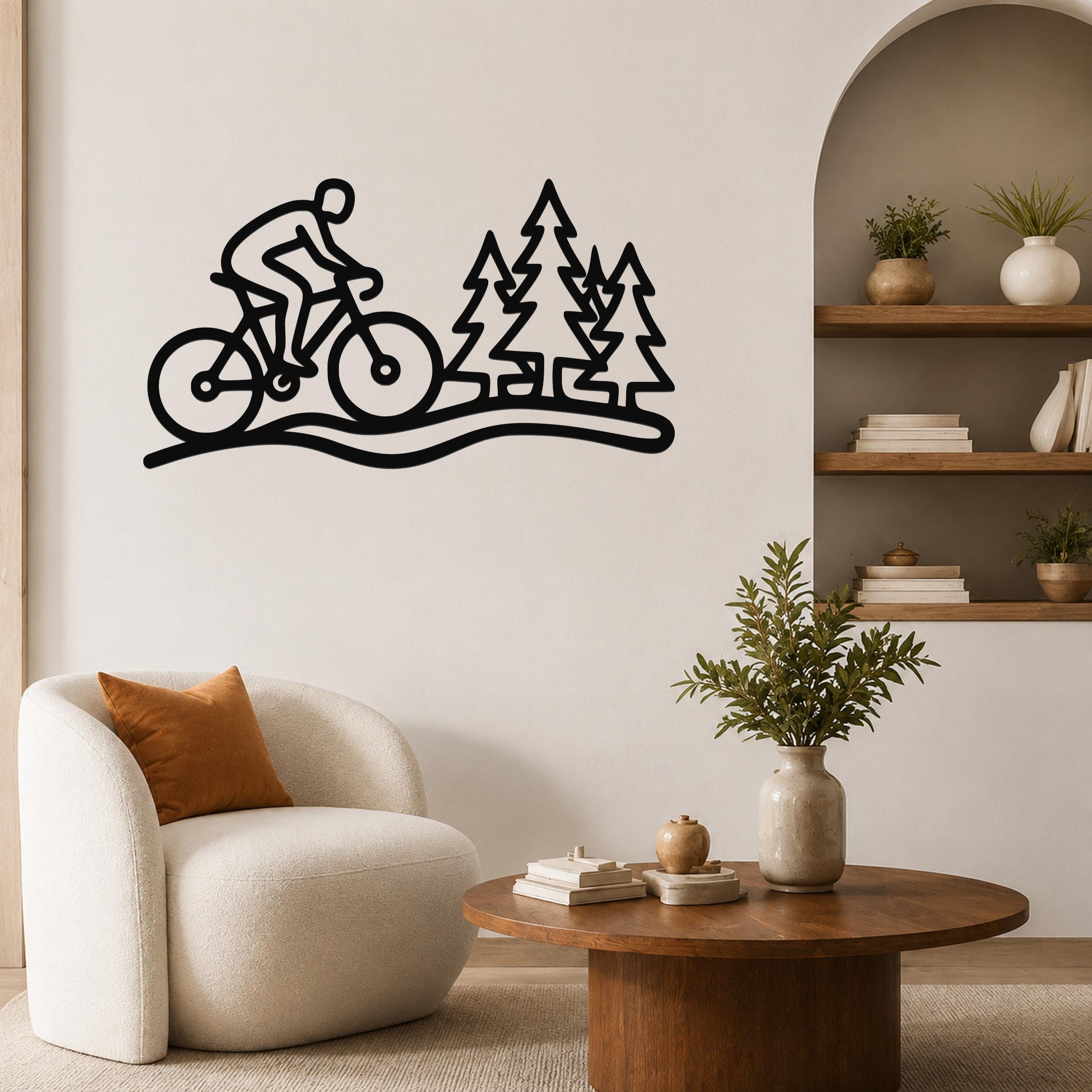 Black metal wall art silhouette of a cyclist on a trail with pine trees, mounted on a living room wall above a white armchair.
