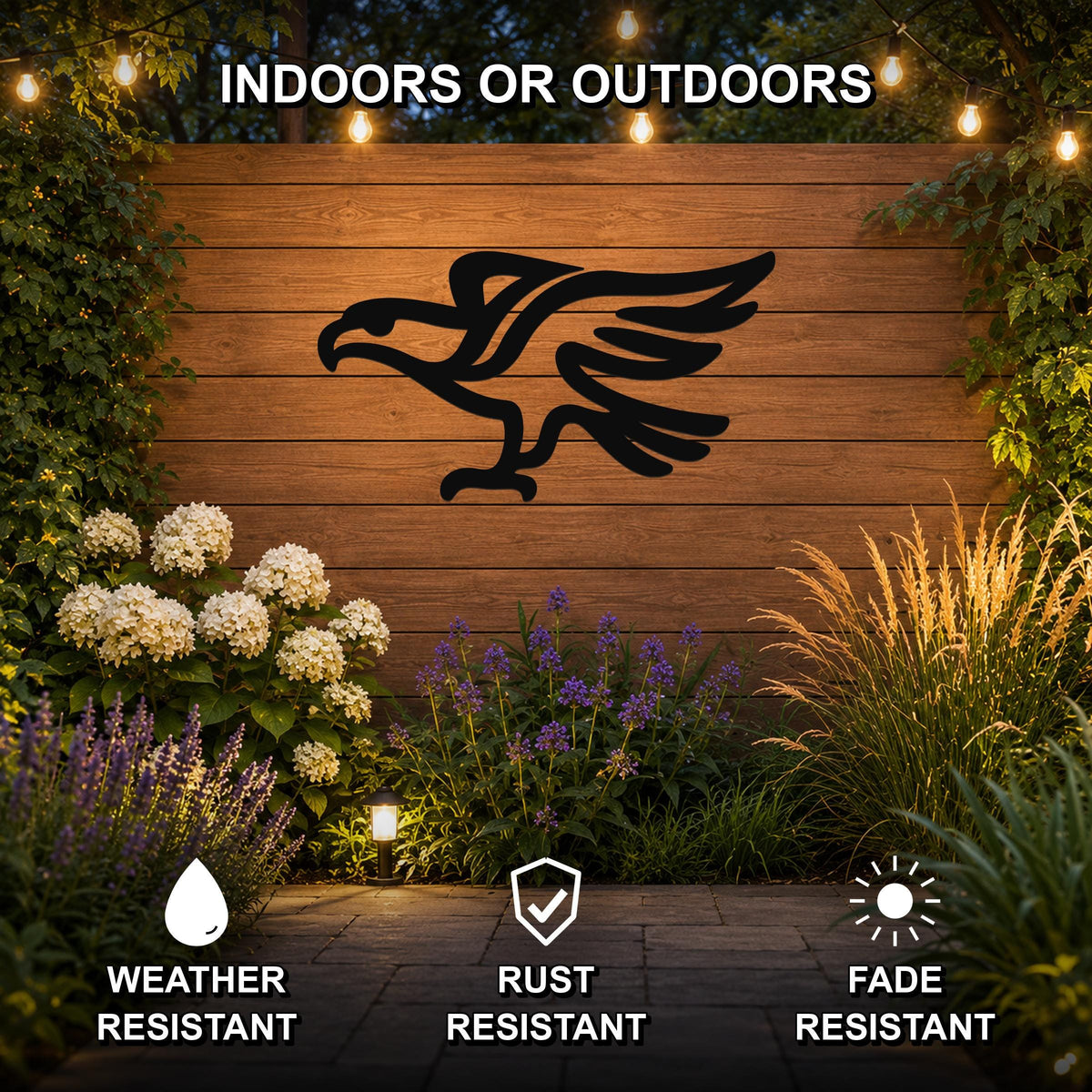 Black metal eagle silhouette wall art mounted on a wooden outdoor wall, displayed above a garden bed with flowers and string lights, weather resistant, rust resistant, and fade resistant.