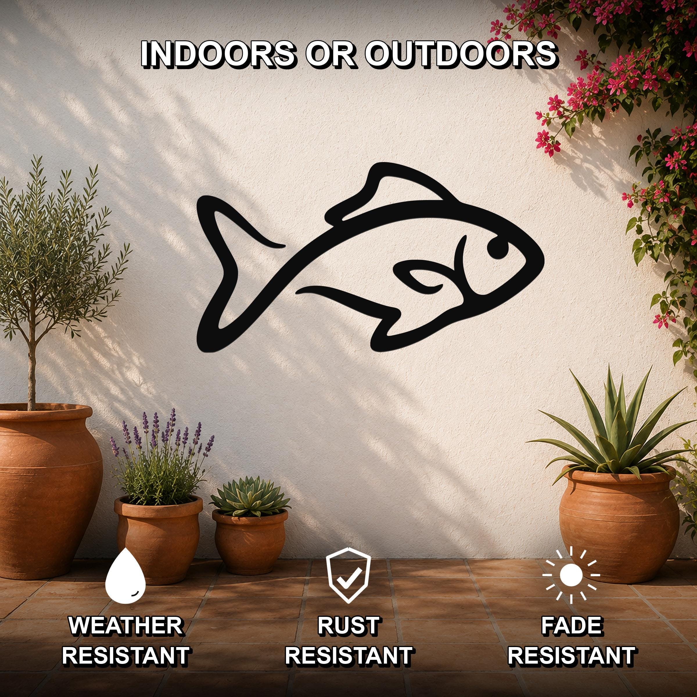 Black metal fish silhouette wall art mounted on a white outdoor wall, surrounded by potted plants and flowering vines, with text indicating it is weather, rust, and fade resistant.