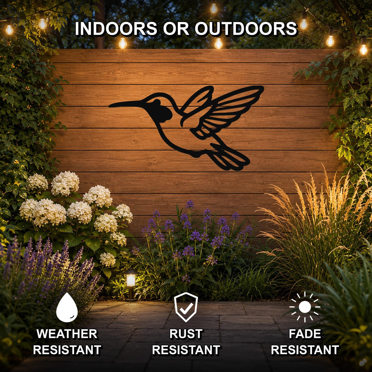 Black metal hummingbird wall art with wing and tail cutouts, mounted on a wooden fence in a garden with string lights and plants.