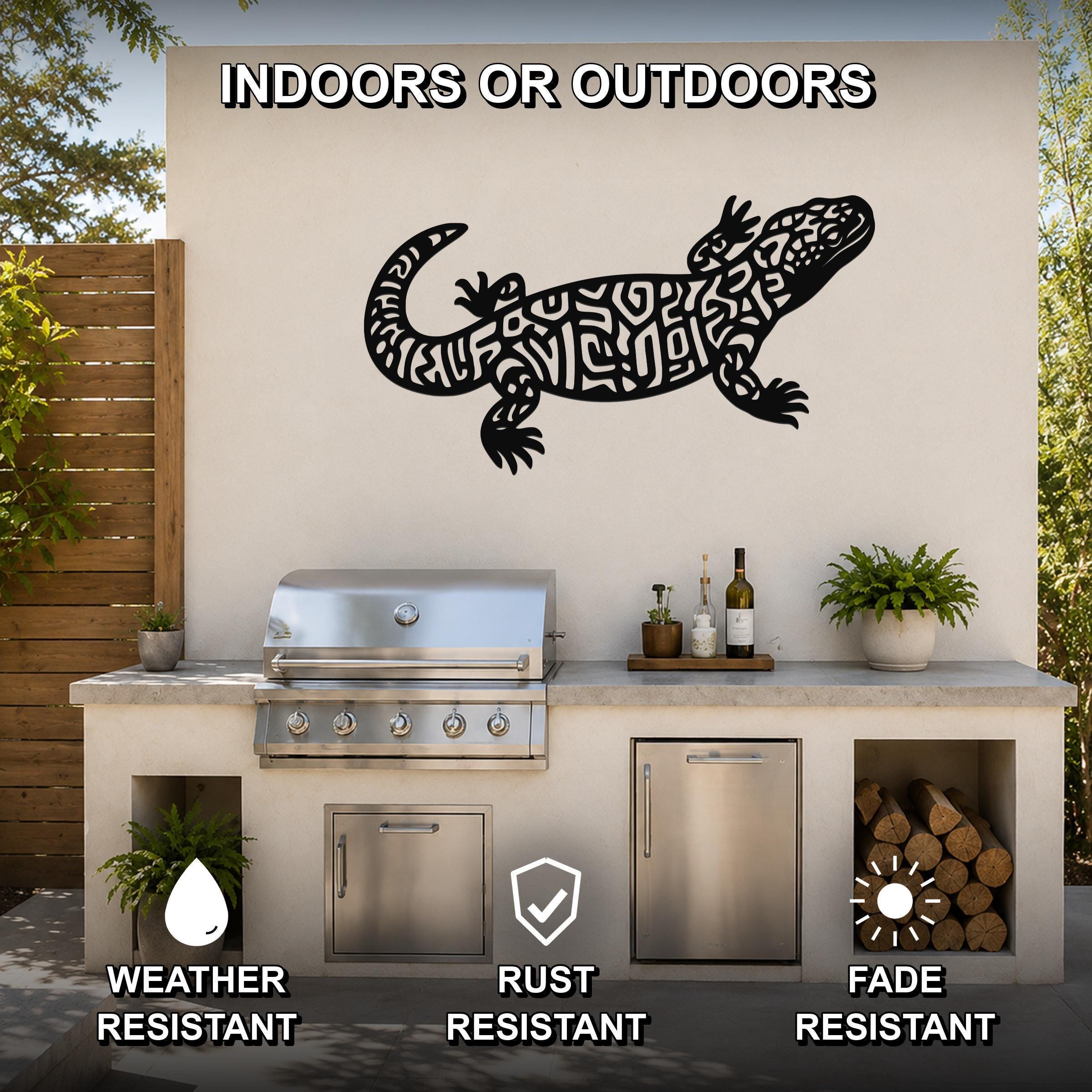 Black metal Gila monster wall art with intricate cutout patterns, mounted on an outdoor kitchen wall above a stainless steel grill.