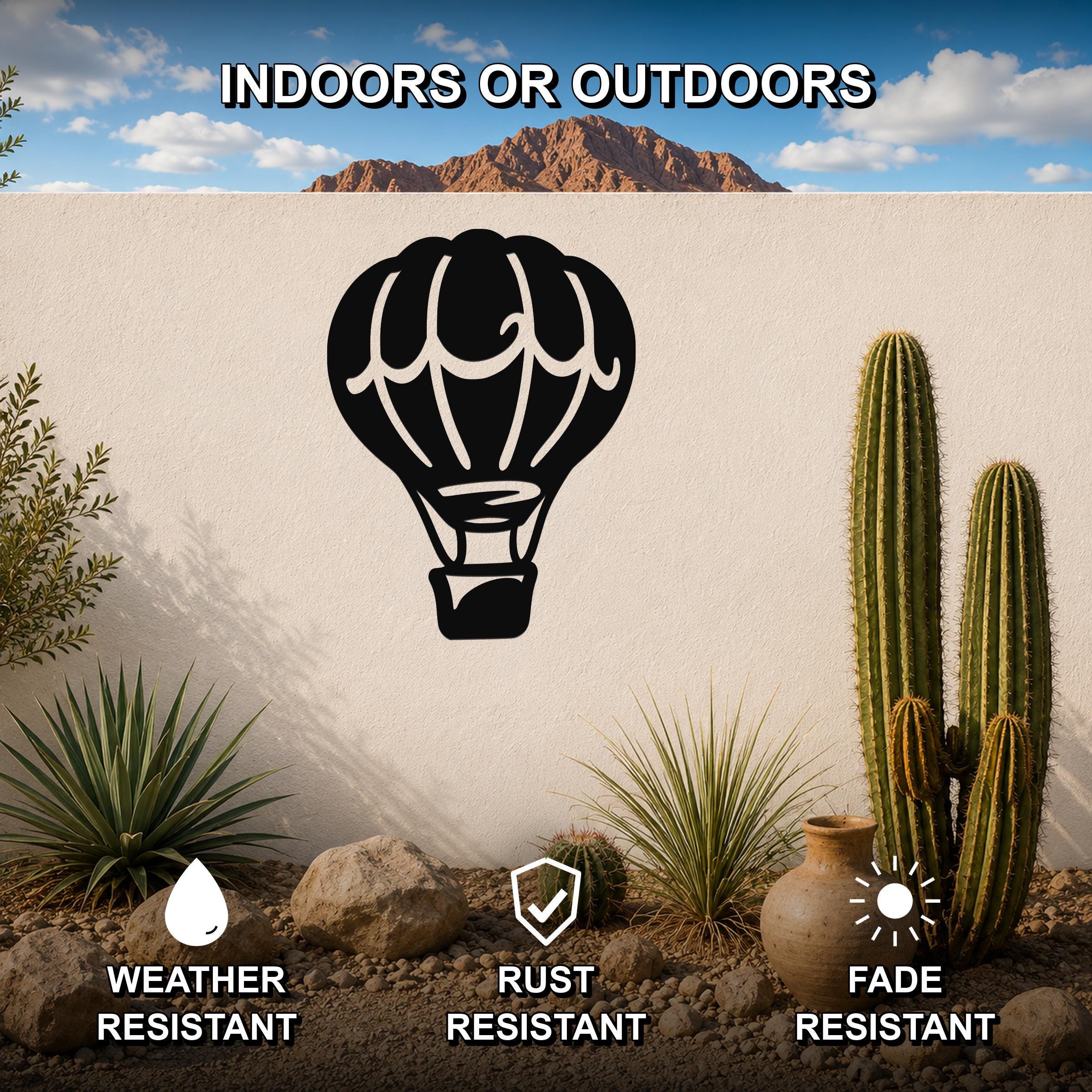 Black metal hot air balloon wall art with internal cutout details, mounted on an outdoor wall in a desert garden setting with cacti and rocks.