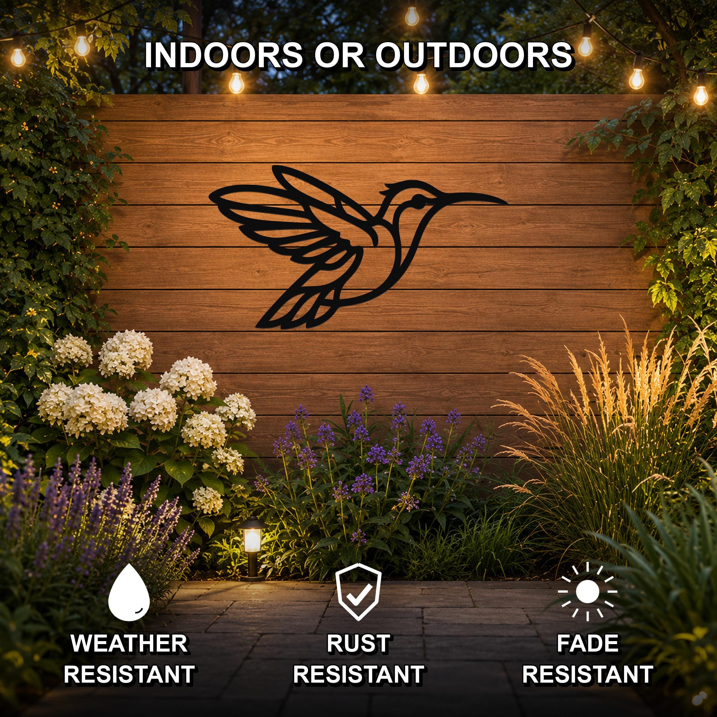 Black metal hummingbird wall art with motion-line cutouts, mounted on a wooden outdoor wall above a garden bed with flowers and string lights.