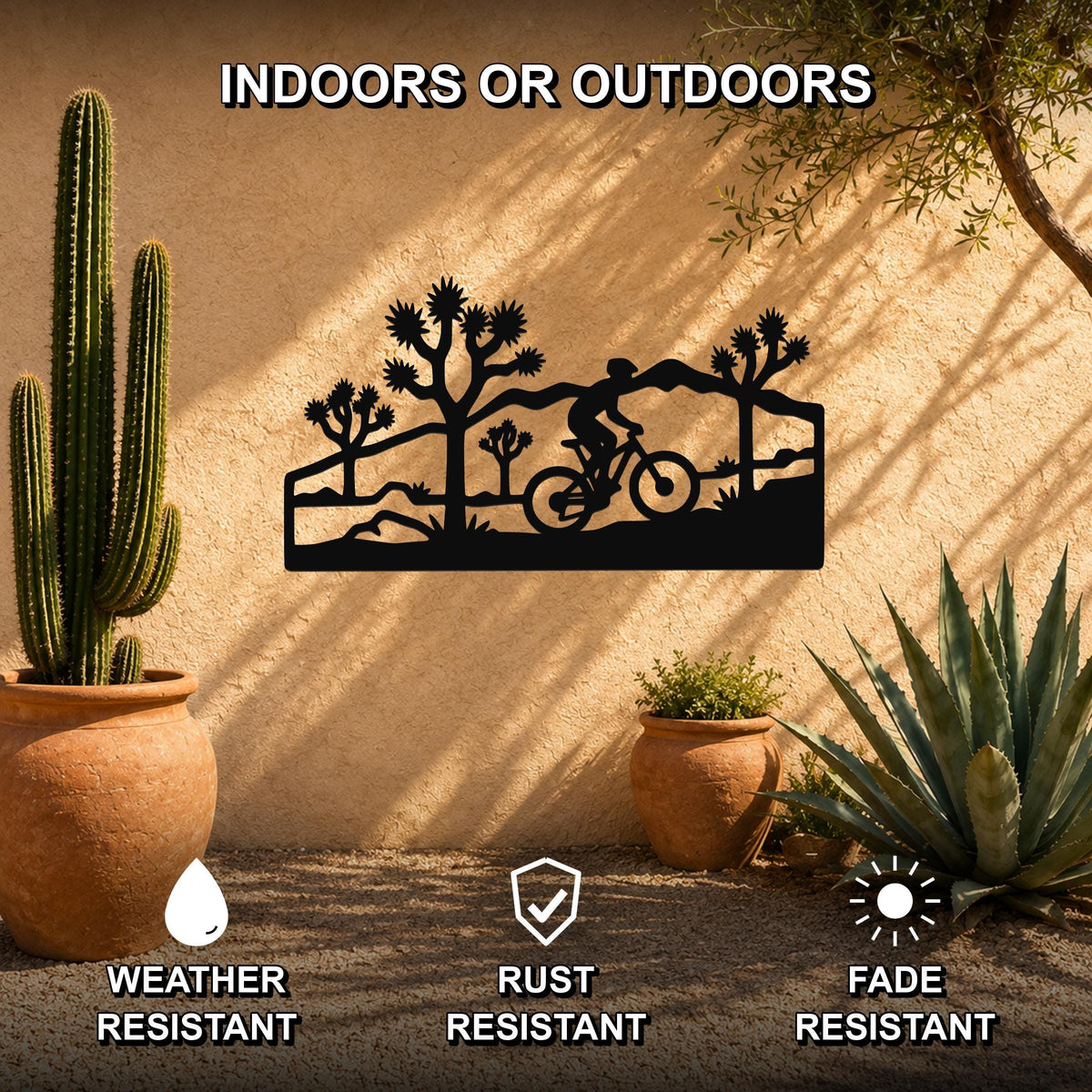 Black metal wall art featuring a cyclist silhouette among Joshua trees, mounted on an outdoor stucco wall with potted cacti and succulents.