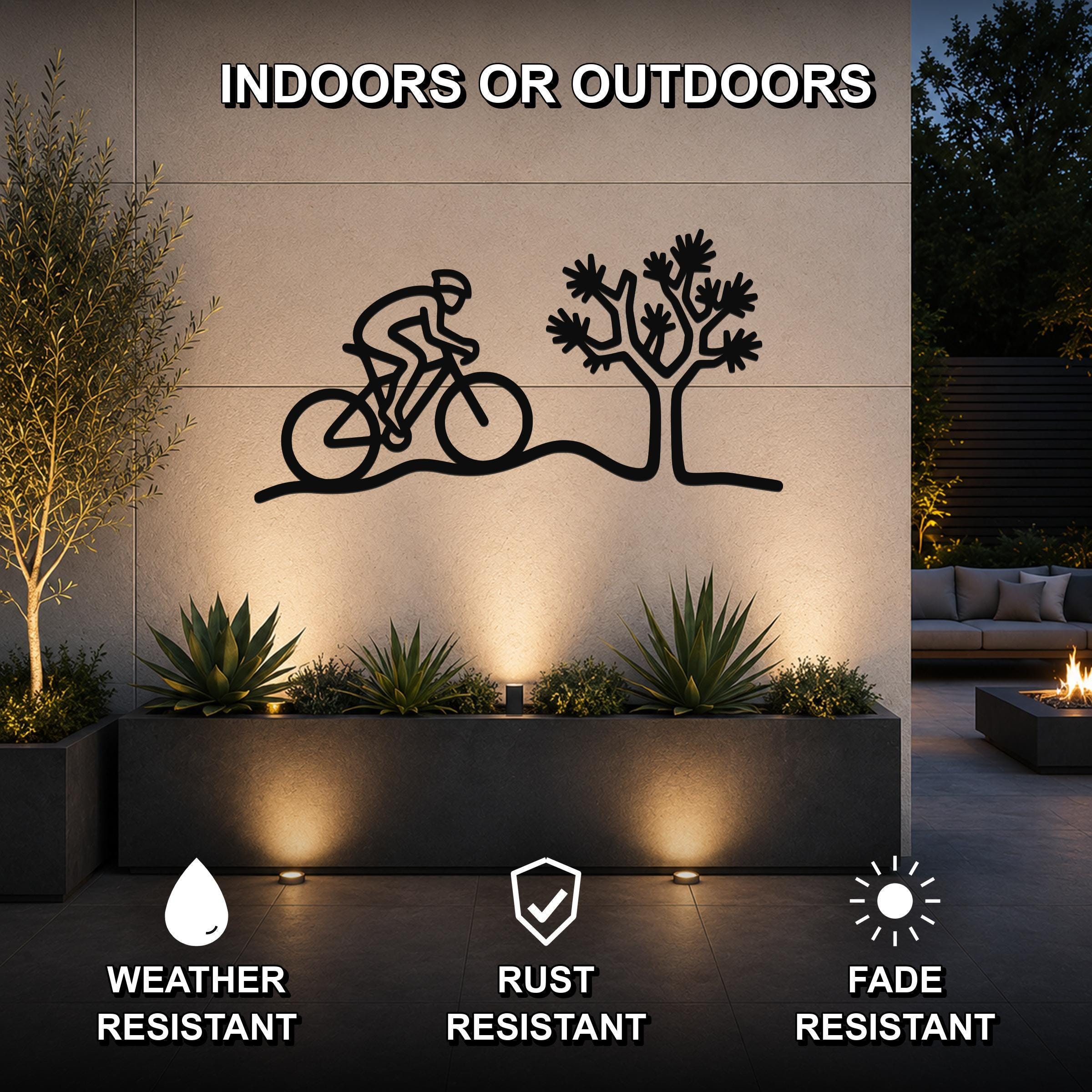 Black metal wall art featuring a cyclist silhouette and a Joshua tree, mounted on an outdoor wall above a planter in a patio setting.