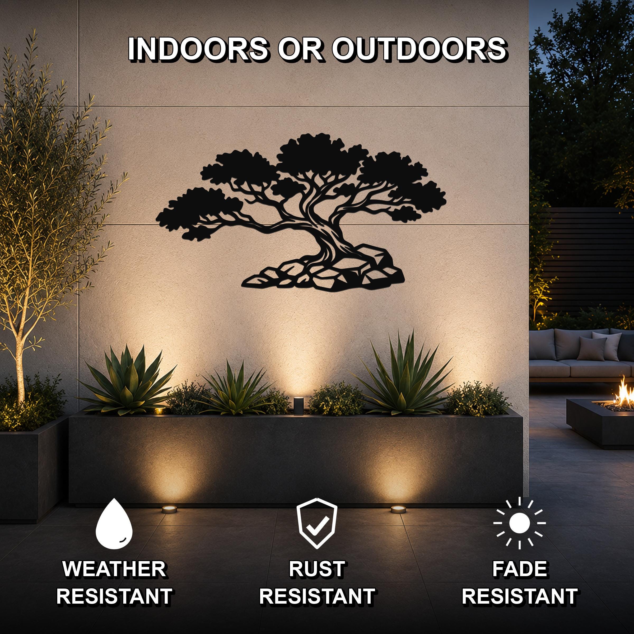 Black metal wall art of a juniper tree silhouette with rocky base, mounted on an outdoor wall above a planter in a patio setting.