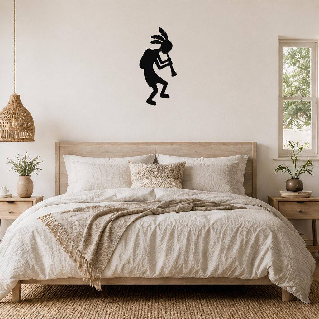 Black metal Kokopelli wall art silhouette playing a flute, mounted on a wall above a bed in a neutral bedroom.
