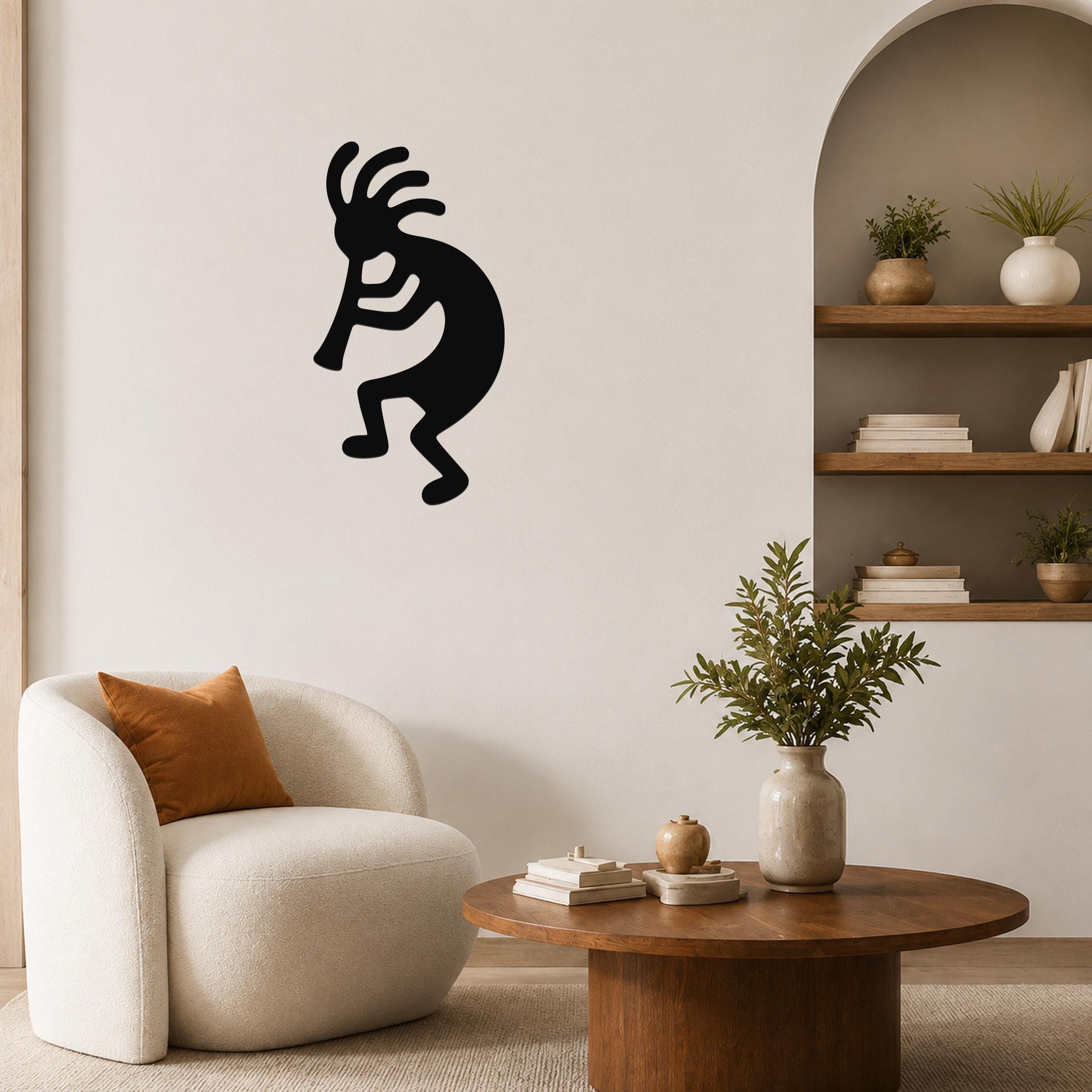 Black metal Kokopelli wall art silhouette with flute player design, mounted on a living room wall above a white armchair.