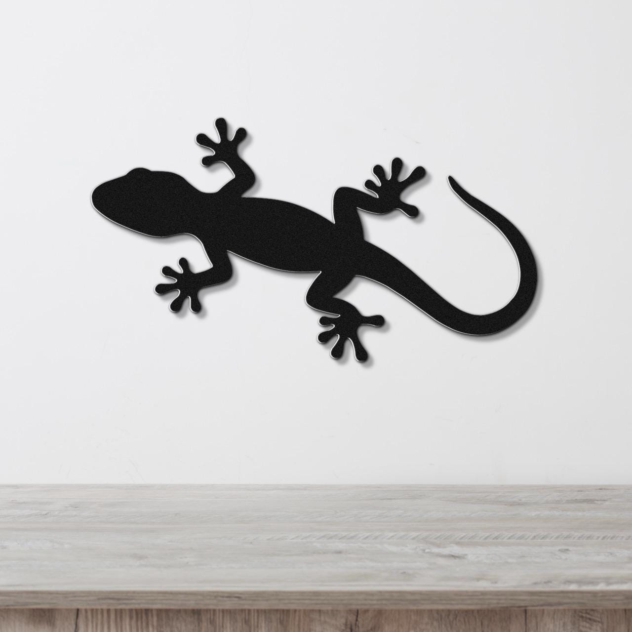 Black metal lizard wall art in a bold silhouette cutout, mounted on a white wall above a light wood surface.