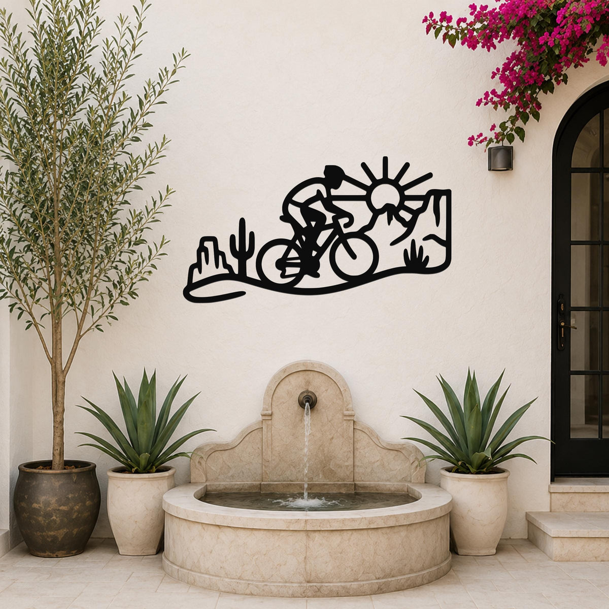 Black metal wall art silhouette of a cyclist on a desert trail with cacti and a sun, mounted on a white outdoor wall above a stone fountain.