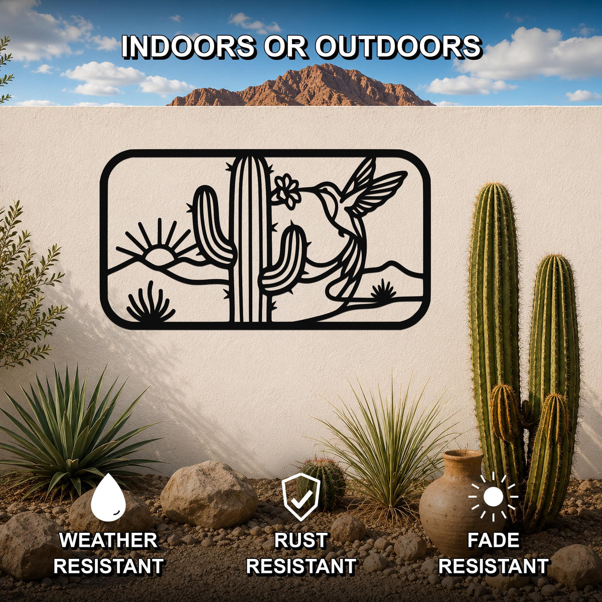Black metal wall art with a desert scene silhouette featuring a saguaro cactus, hummingbird, and sun, mounted on an outdoor wall above a rock and cactus garden.