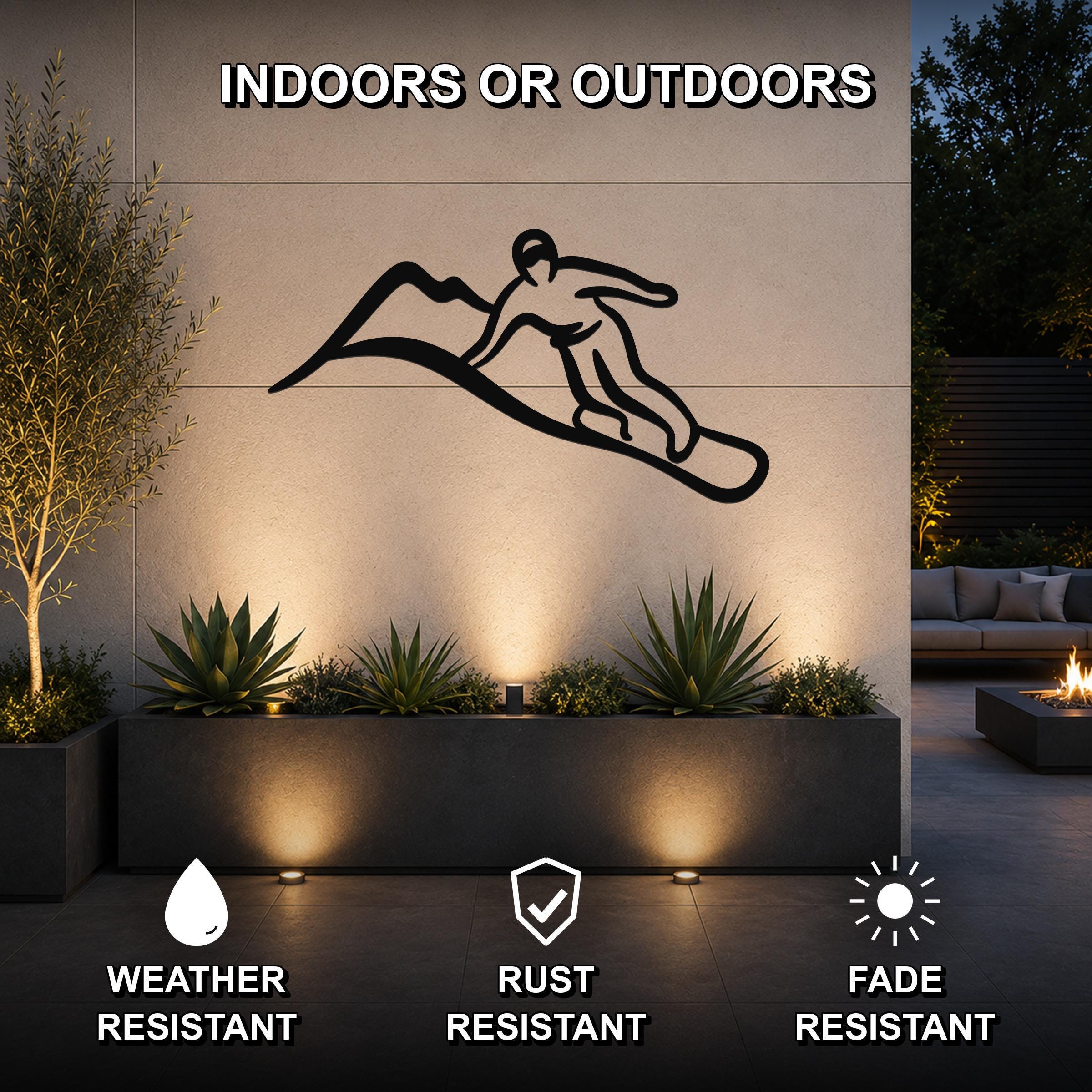 Black metal wall art silhouette of a snowboarder on a mountain, mounted on an outdoor wall above a planter with plants and uplighting.