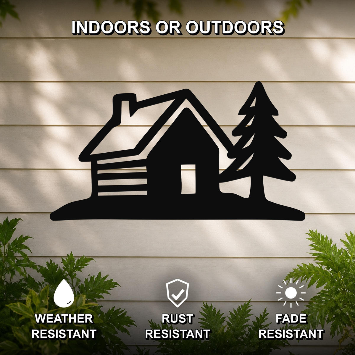 Black metal wall art featuring a cabin and pine tree silhouette, mounted on a light-colored outdoor wall with greenery below.