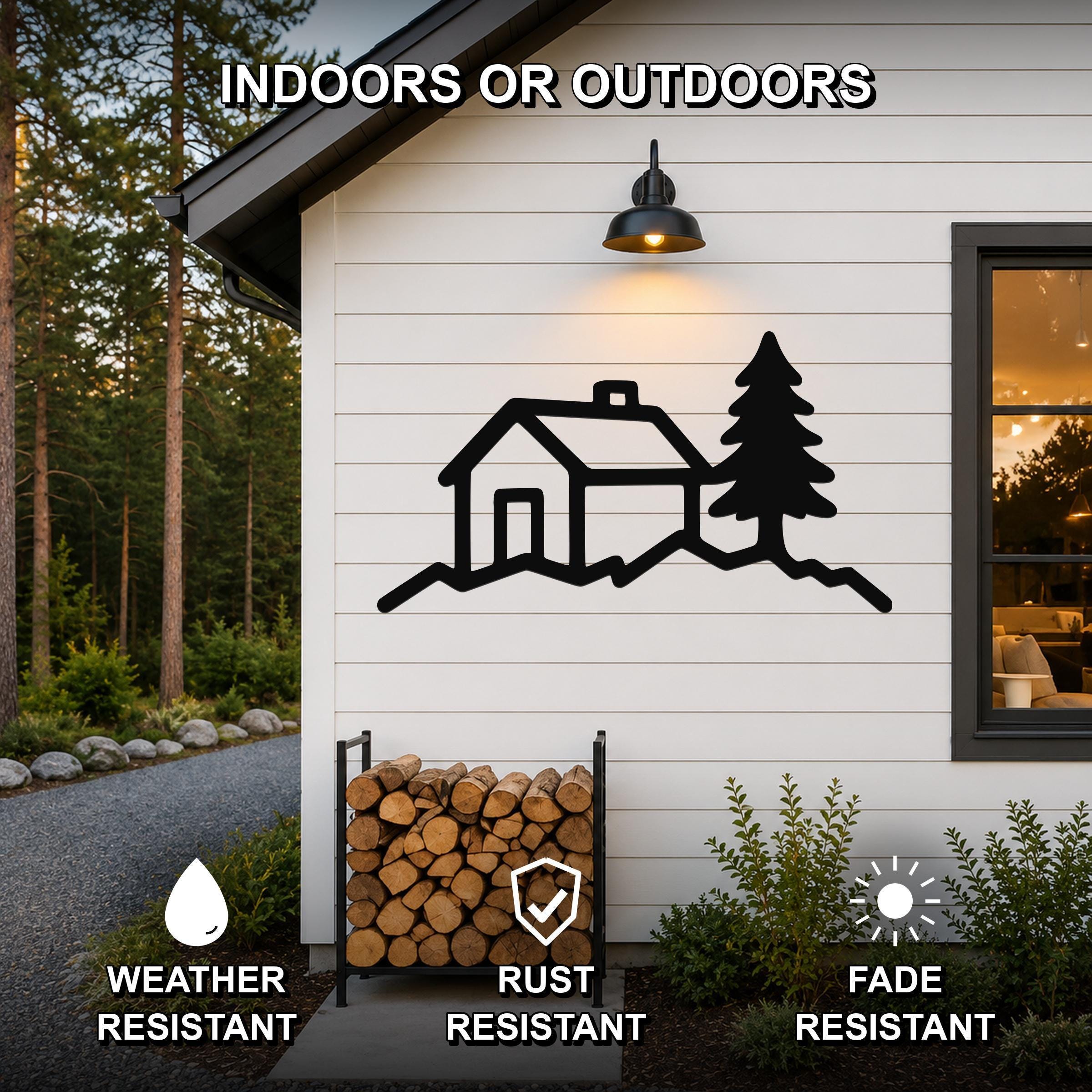 Black metal wall art featuring a cabin and pine tree silhouette, mounted on a white exterior wall above a firewood rack, with text indicating it is weather, rust, and fade resistant.