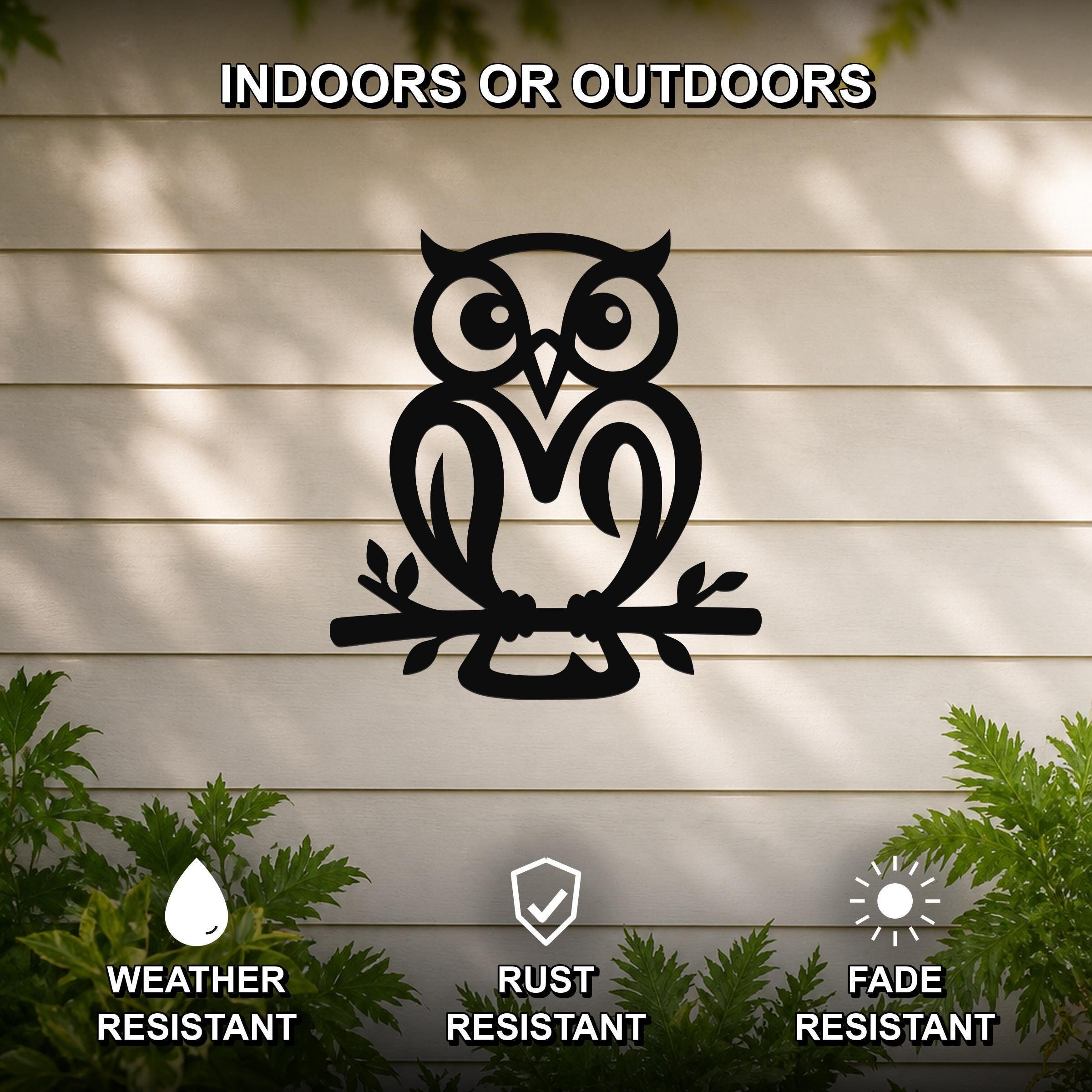 Black metal owl wall art cutout perched on a branch, mounted on a beige outdoor wall with greenery, weather, rust, and fade resistant.