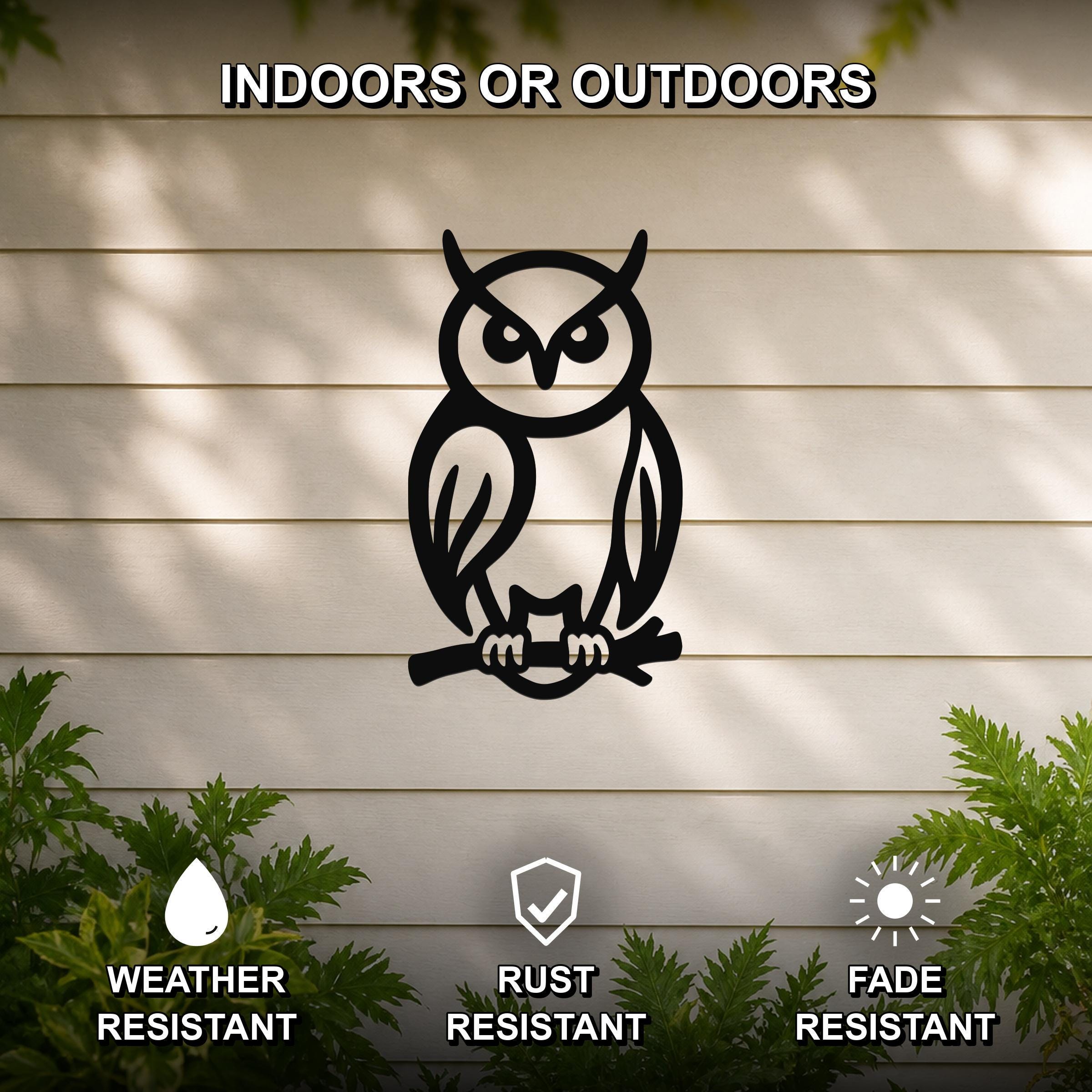 Black metal owl wall art in a silhouette style, perched on a branch, mounted on a light-colored outdoor wall with greenery below.