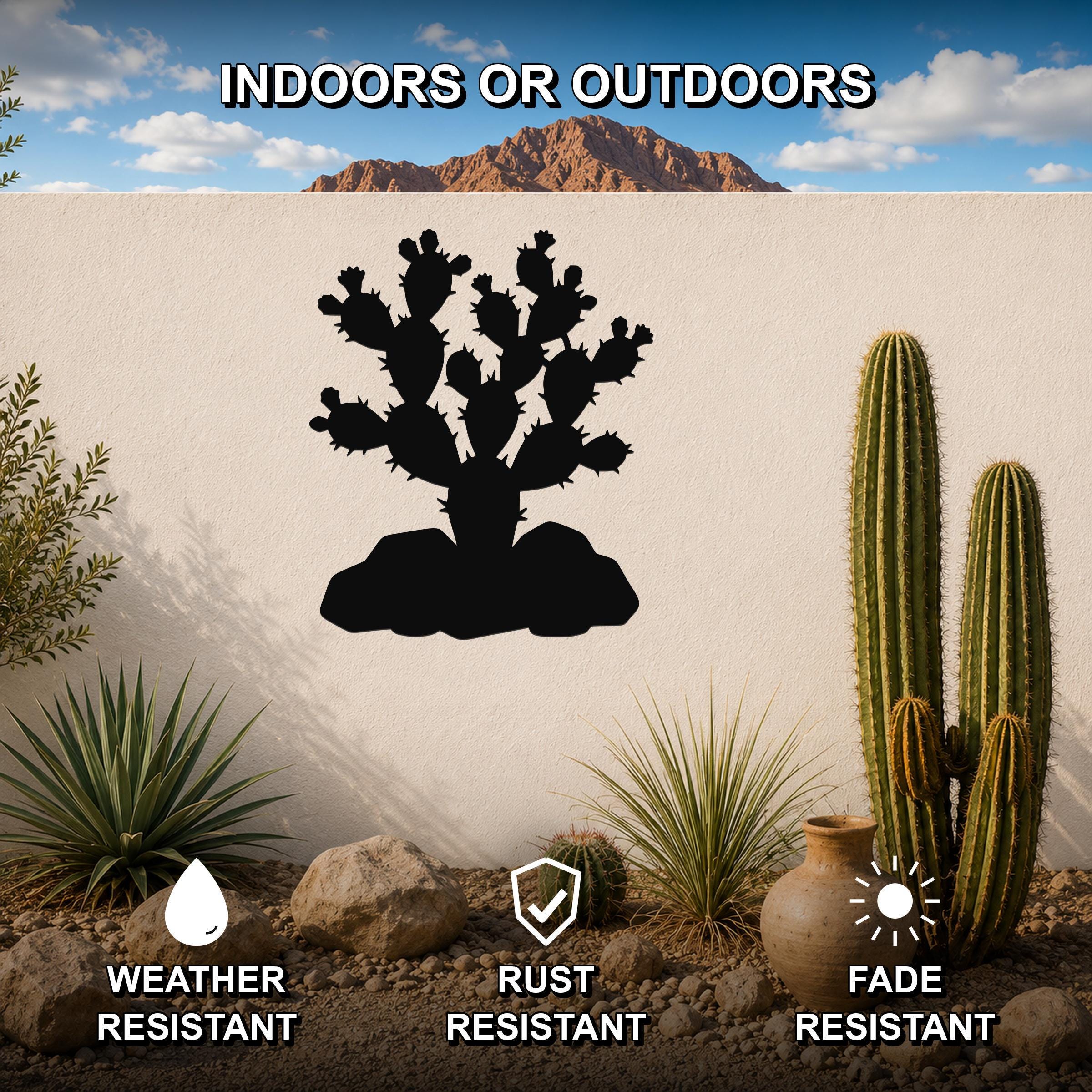 Black metal prickly pear cactus wall art silhouette mounted on a white outdoor wall, surrounded by desert plants and rocks under a blue sky.