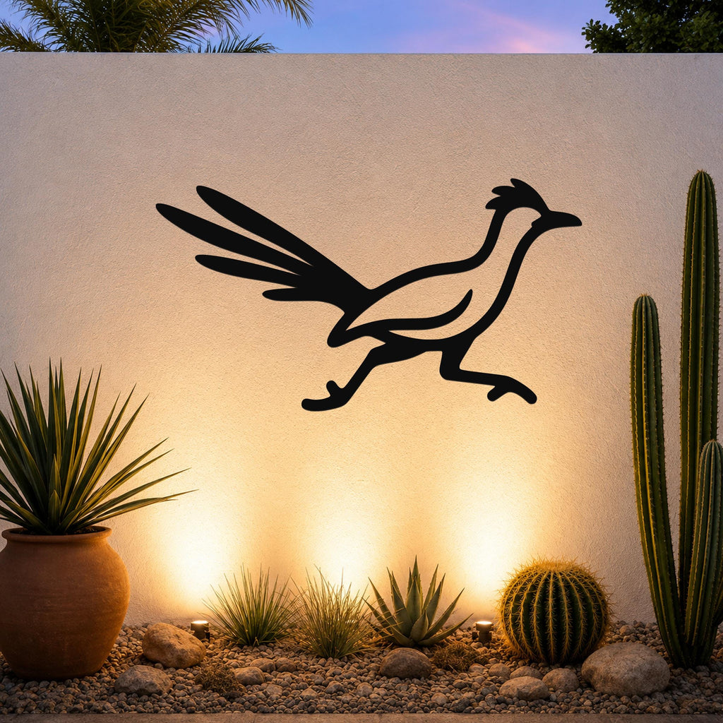 Black metal roadrunner wall art silhouette with spread wings and long tail, mounted on a textured outdoor wall above a desert garden with cacti and warm uplighting.
