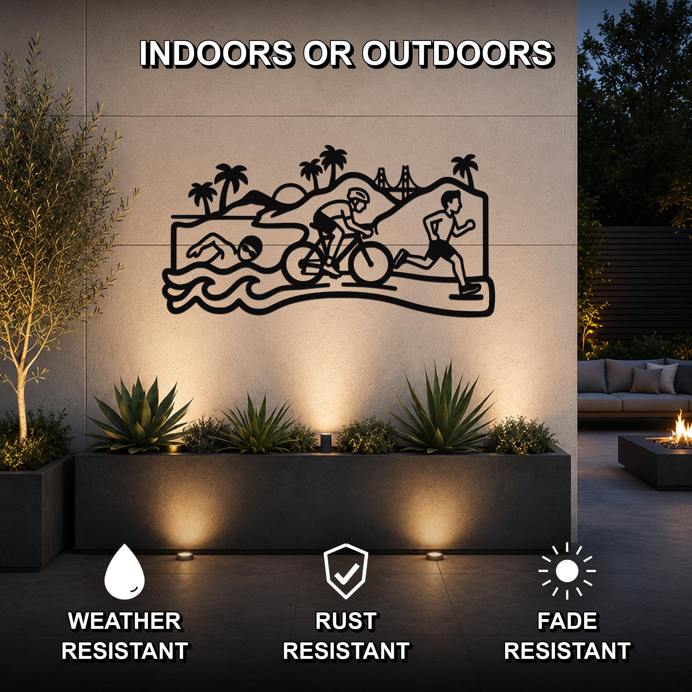 Black metal wall art featuring a triathlon scene with swim, bike, and run silhouettes, mounted on an outdoor patio wall above a planter with plants and uplighting.