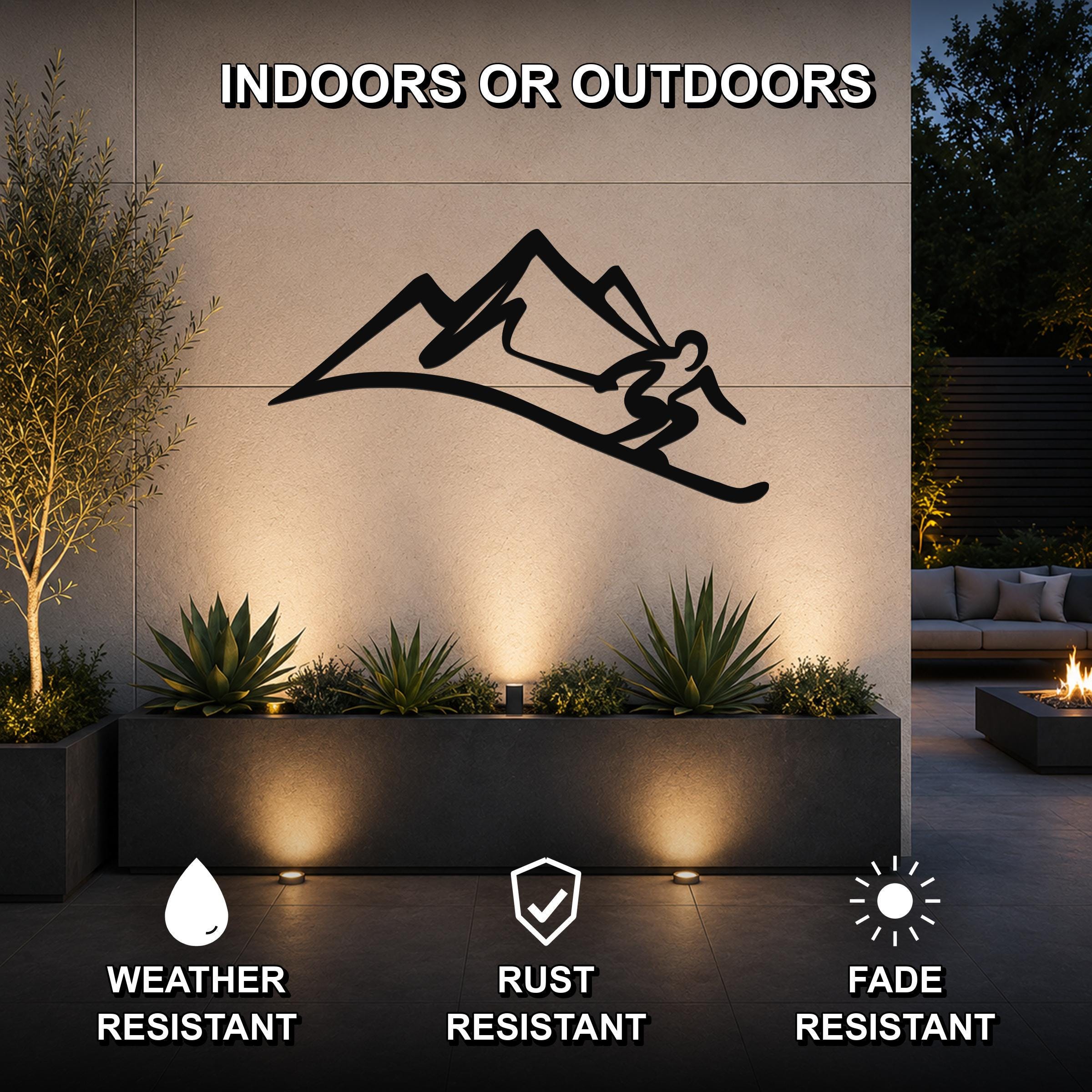 Black metal skier and mountain wall art silhouette mounted on an outdoor patio wall above a planter, with lighting highlighting the scene.
