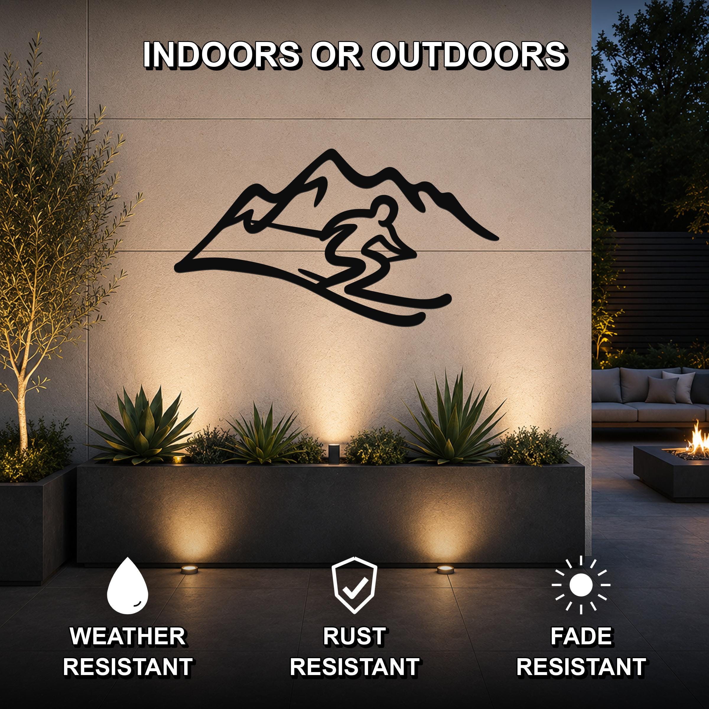 Black metal skier and mountain wall art silhouette mounted on an outdoor patio wall above a planter, with text indicating it is weather, rust, and fade resistant.