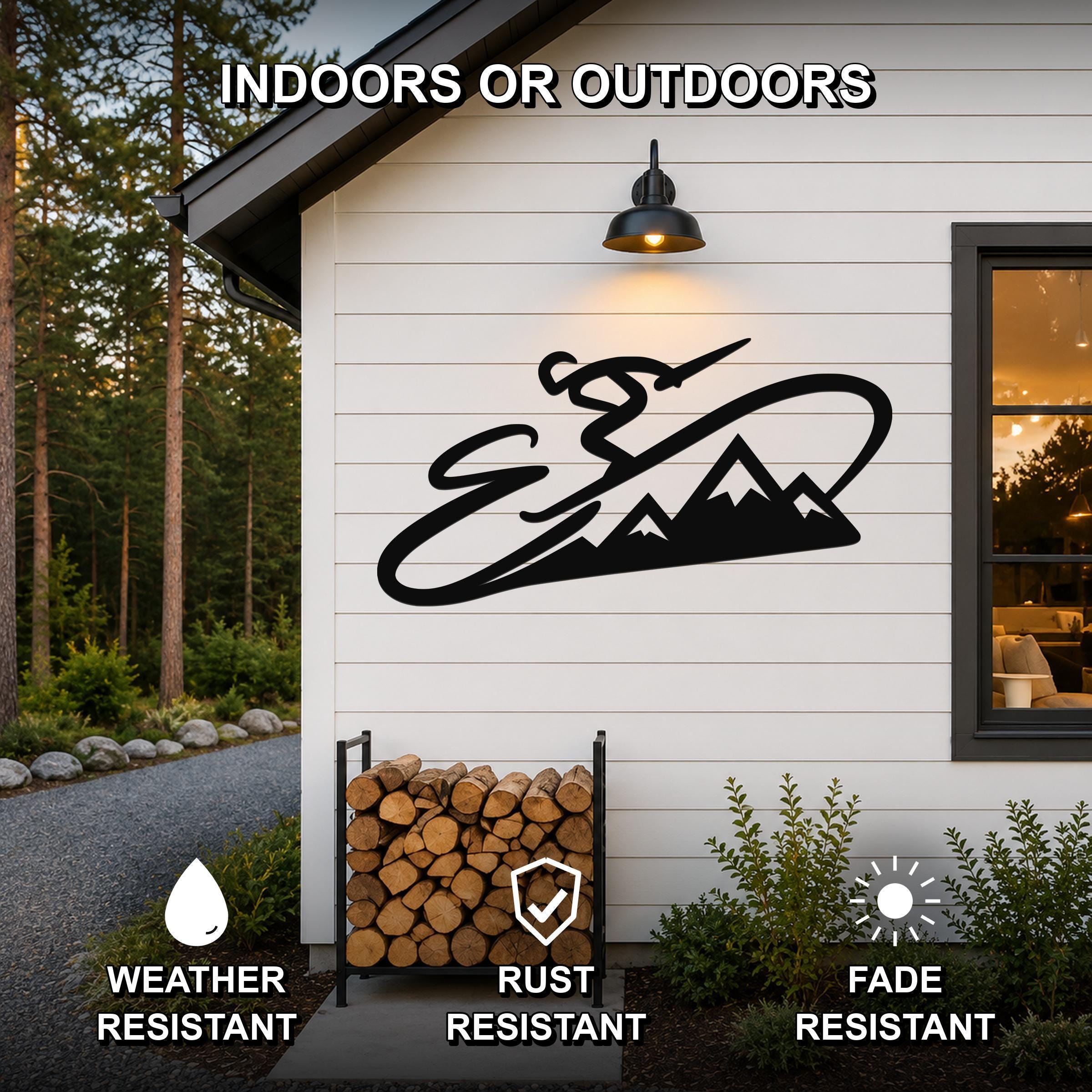 Black metal wall art featuring a skier silhouette and mountain range, mounted on a white exterior wall above a firewood rack, with text indicating it is weather, rust, and fade resistant.