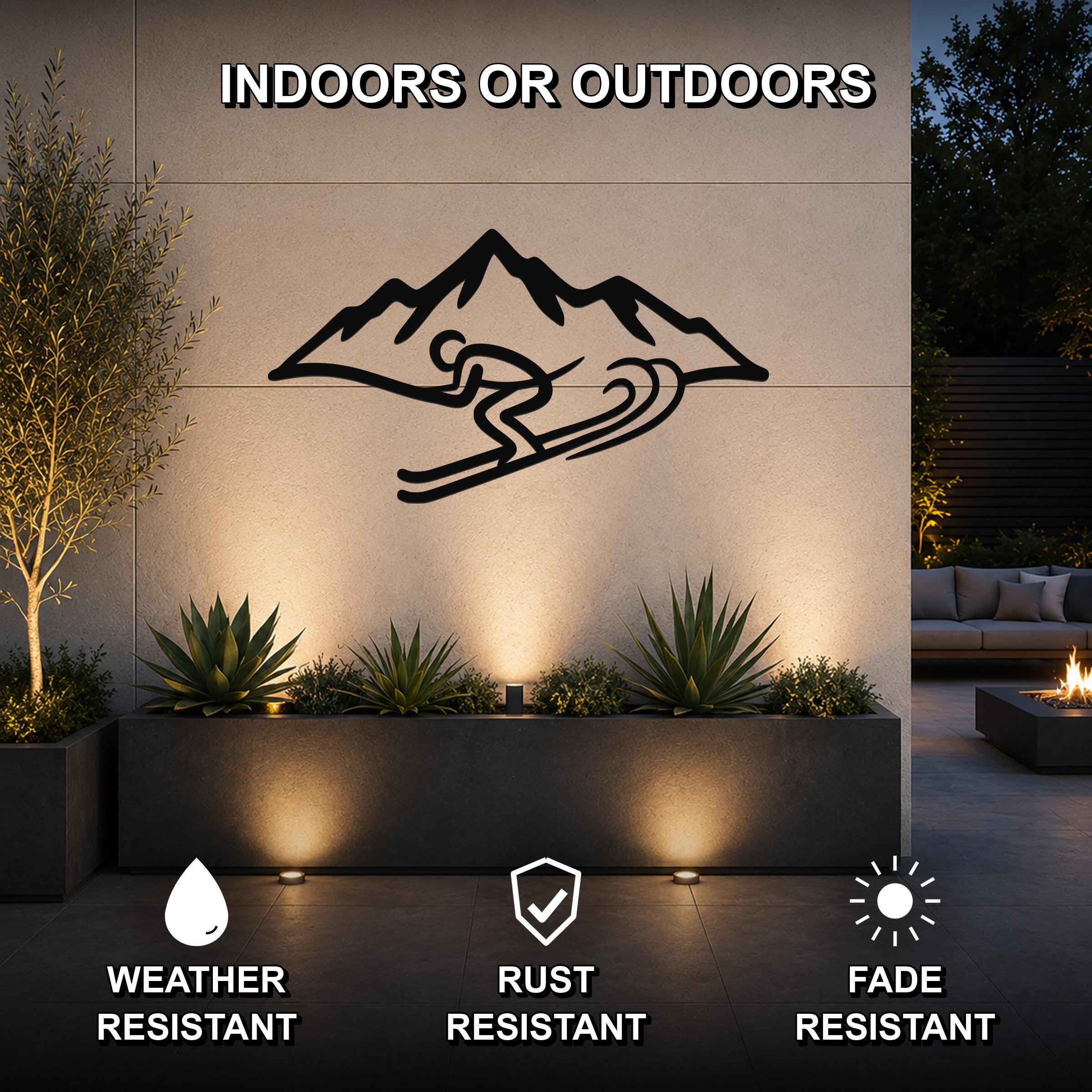 Black metal skier and mountain wall art silhouette mounted on an outdoor patio wall above a planter, with text indicating it is weather, rust, and fade resistant for indoor or outdoor use.