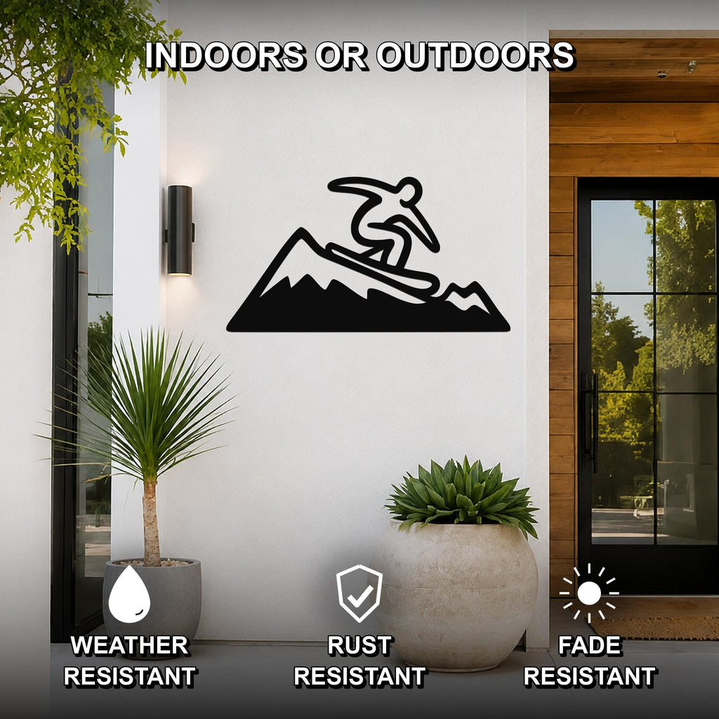 Black metal wall art silhouette of a snowboarder on a mountain, mounted on a white exterior wall beside a glass door and potted plants.