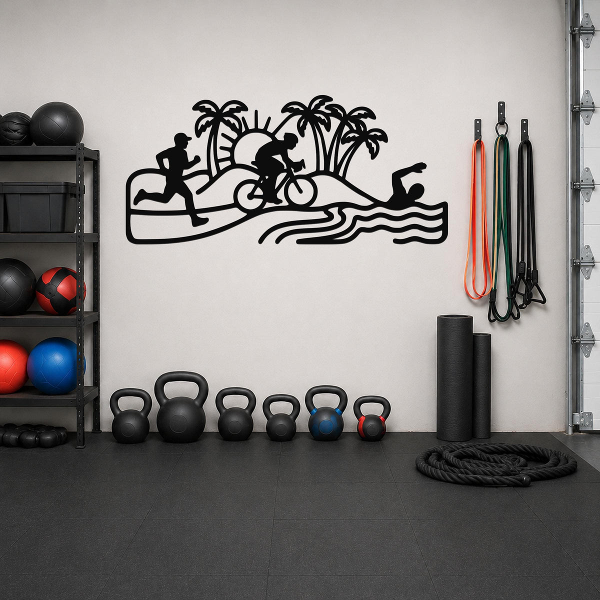 Black metal wall art cutout of a triathlon scene with swim, bike, and run silhouettes, palm trees, and waves, mounted on a gym wall above a row of kettlebells.