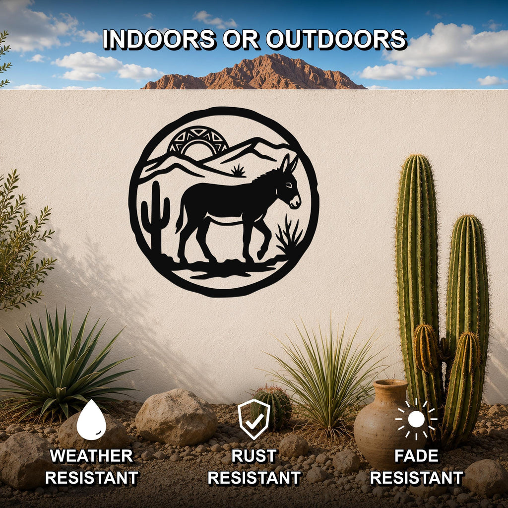 Black metal wall art featuring a burro silhouette within a circular desert scene with cacti and mountains, mounted on an outdoor wall with desert plants and rocks below.