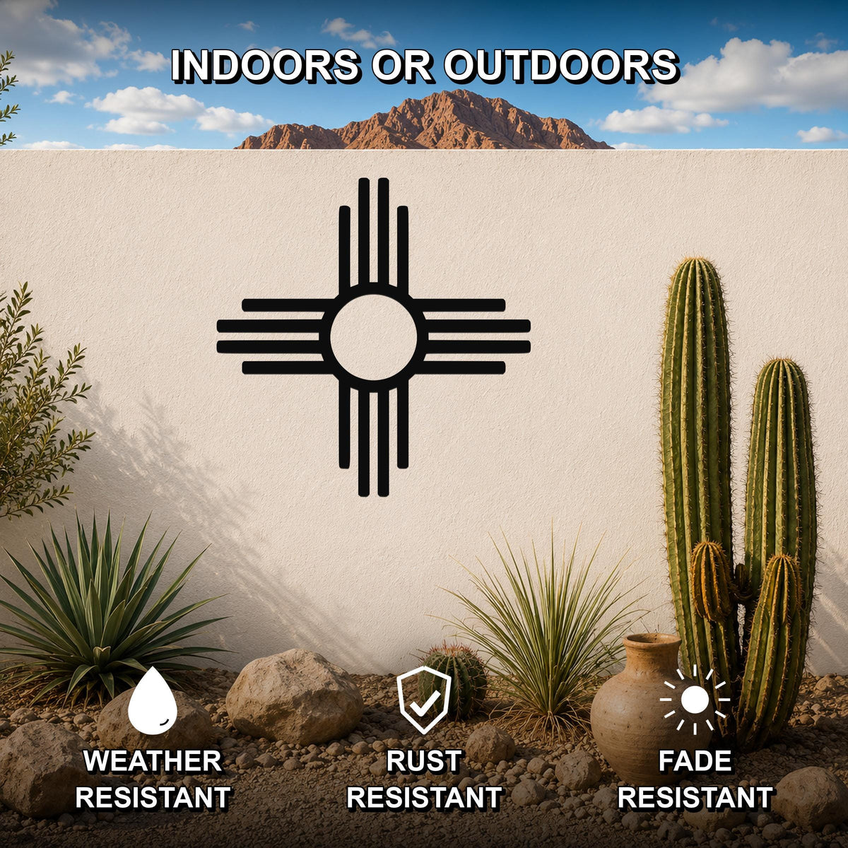 Black metal Zia Sun symbol wall art with a bold circular design and radiating lines, mounted on a white outdoor wall in a desert garden setting with cacti and rocks.