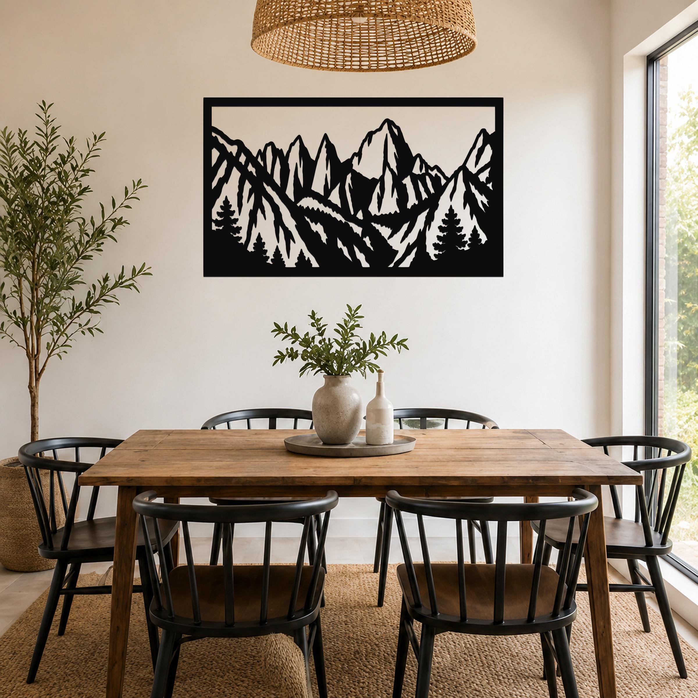 Black metal wall art with a mountain range and tree silhouette cutout, mounted above a wooden dining table in a bright dining room.