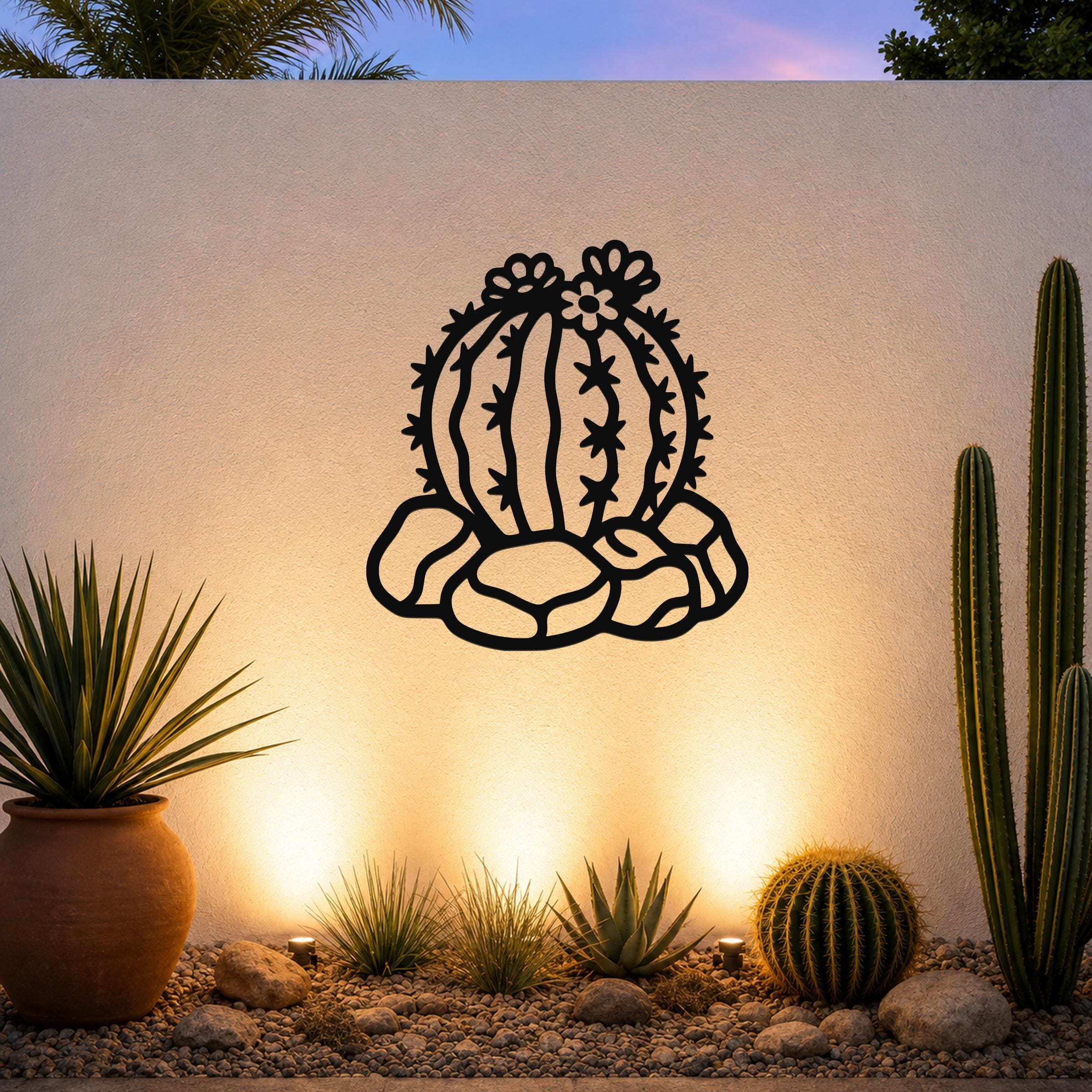 Black metal barrel cactus wall art with flower and spine cutouts, mounted on a white outdoor wall above a desert garden with potted plants and ground lights.