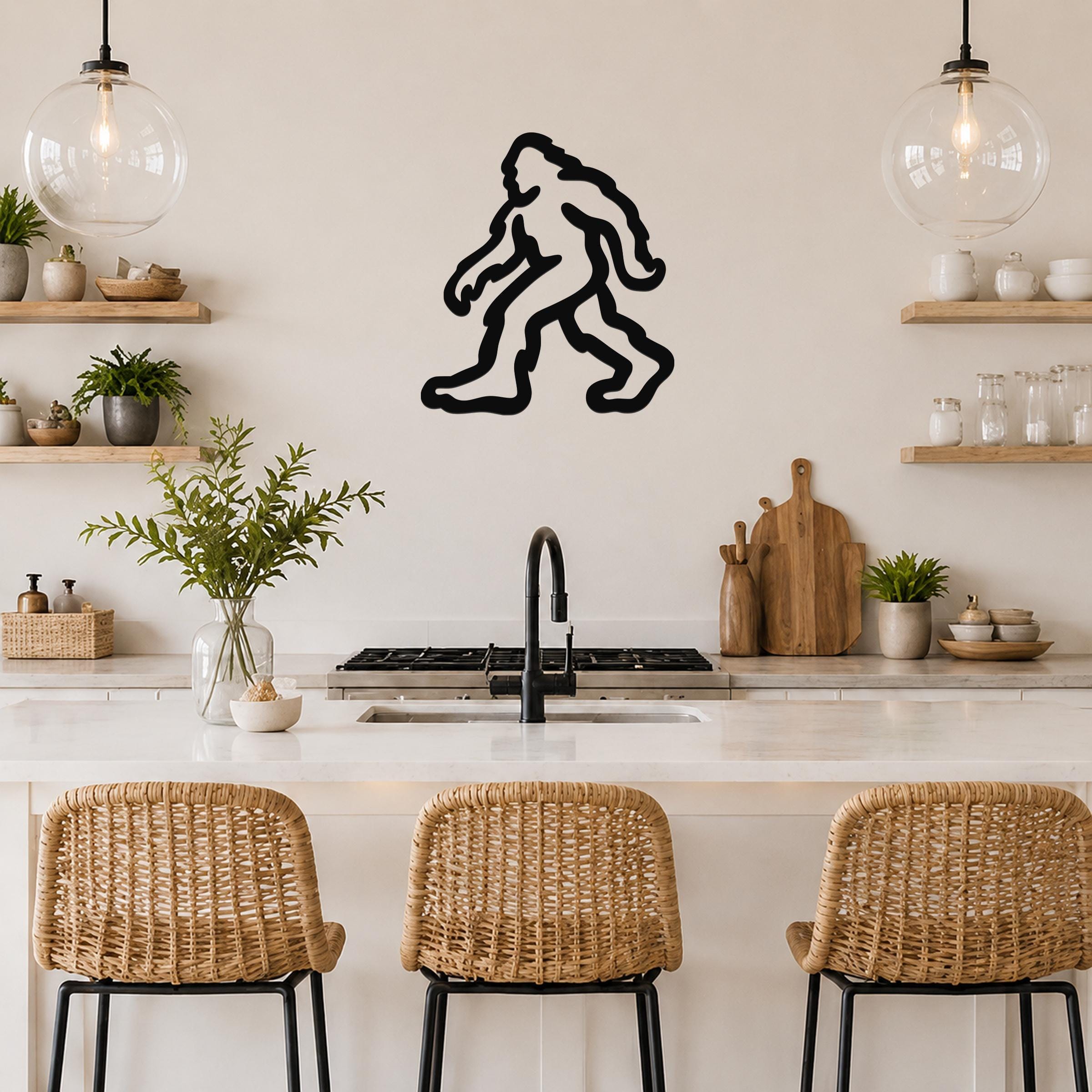 Black metal Bigfoot silhouette wall art mounted on a kitchen wall above a counter, with wicker bar stools and open shelves in the background.