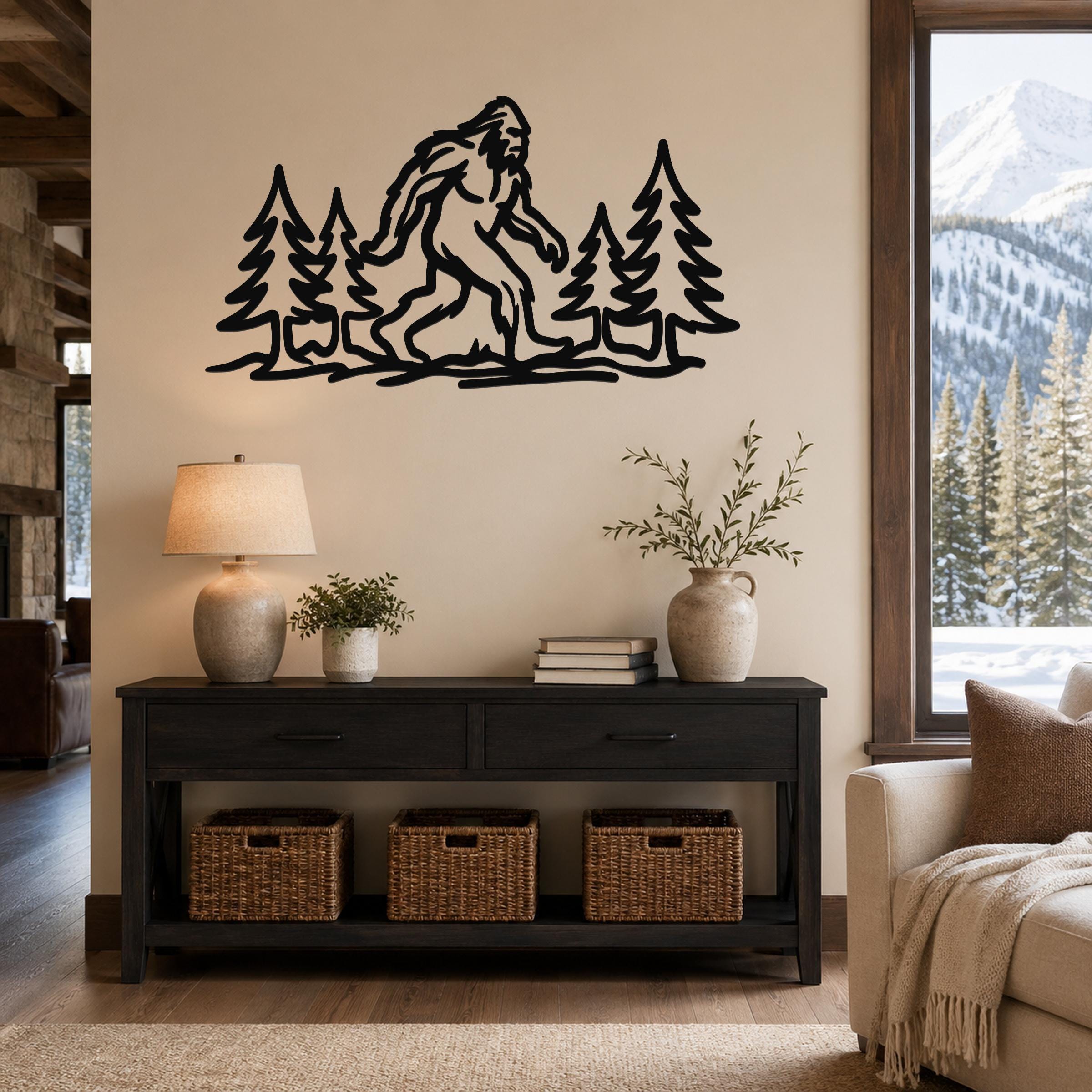 Black metal wall art featuring a Bigfoot silhouette among pine trees, mounted above a dark console table in a living room with a snowy mountain view.