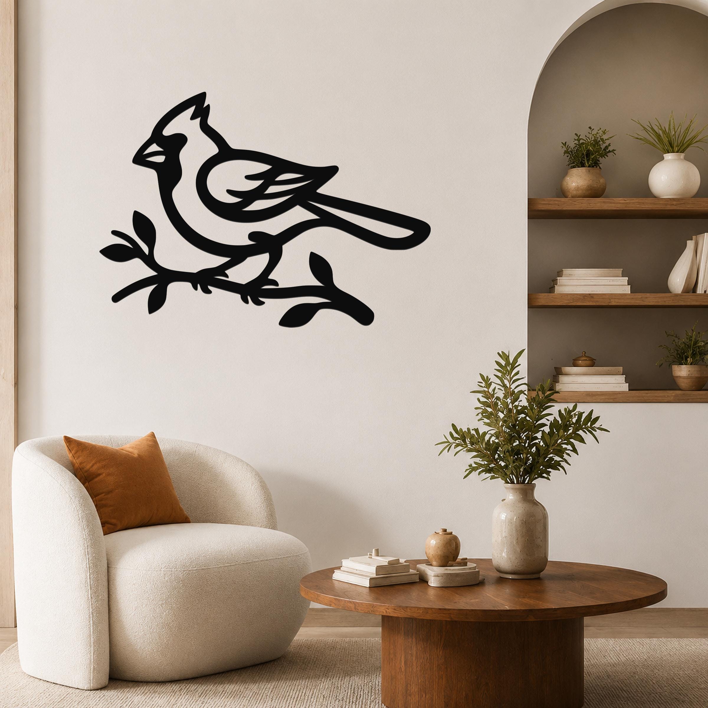 Black metal cardinal wall art in silhouette, perched on a branch, mounted on a living room wall above a white armchair.