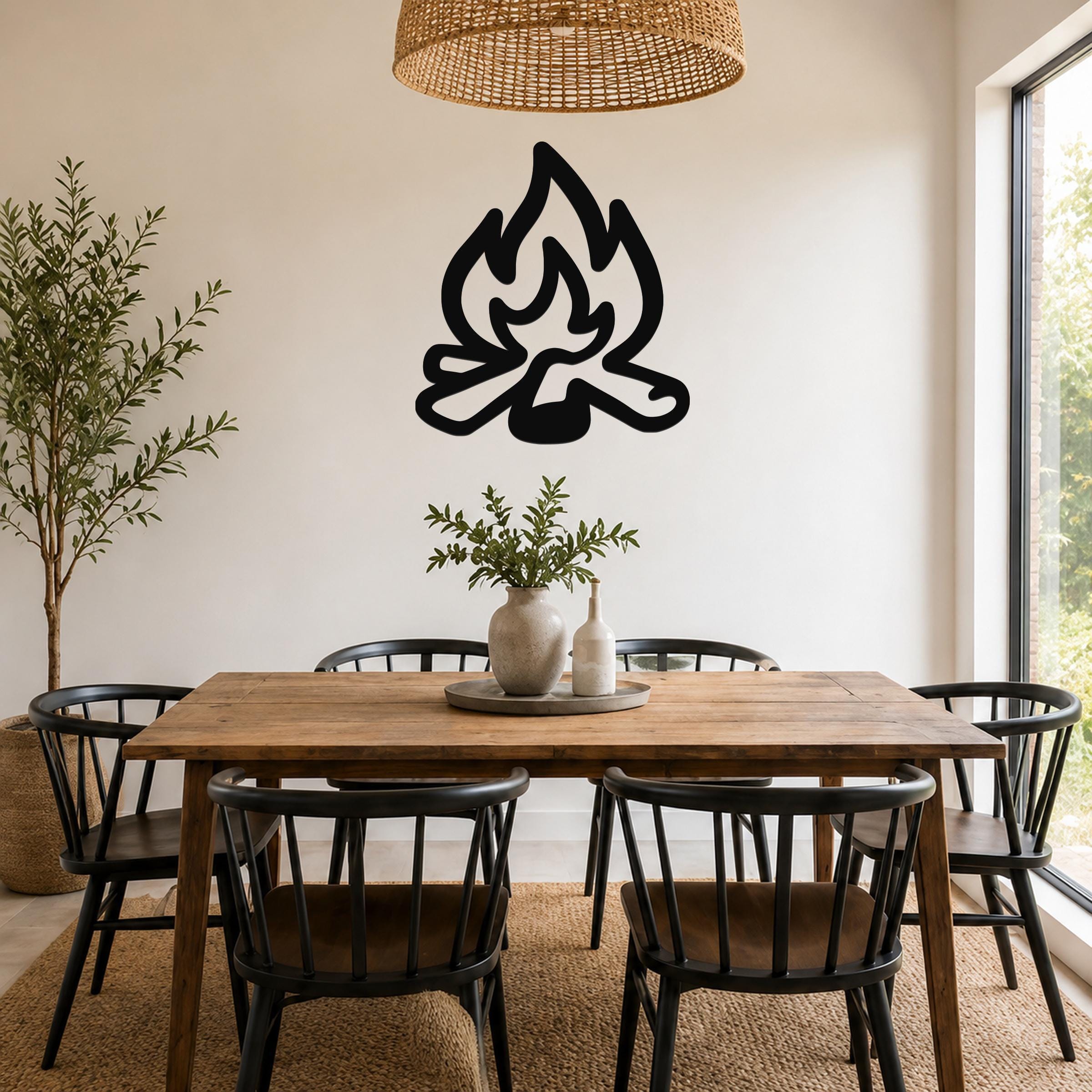 Black metal campfire wall art silhouette with flame and log cutouts, mounted on a white wall above a wooden dining table in a bright dining room.
